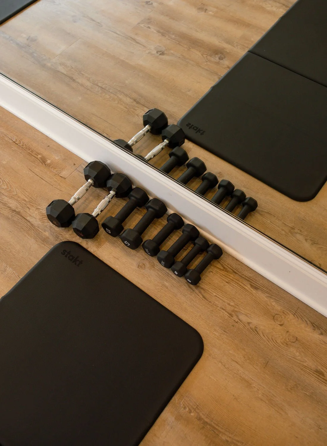 A set of dumbbells and weight plates on a wooden floor next to a mirror and black exercise mats.