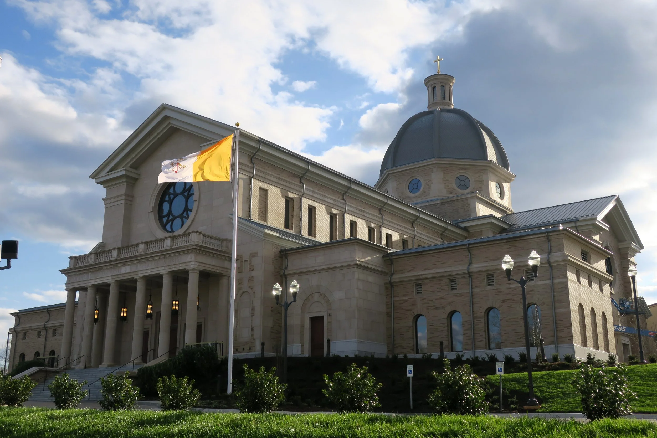 Church building with a domed roof, cross on top, flags, and surrounding greenery under a cloudy sky.