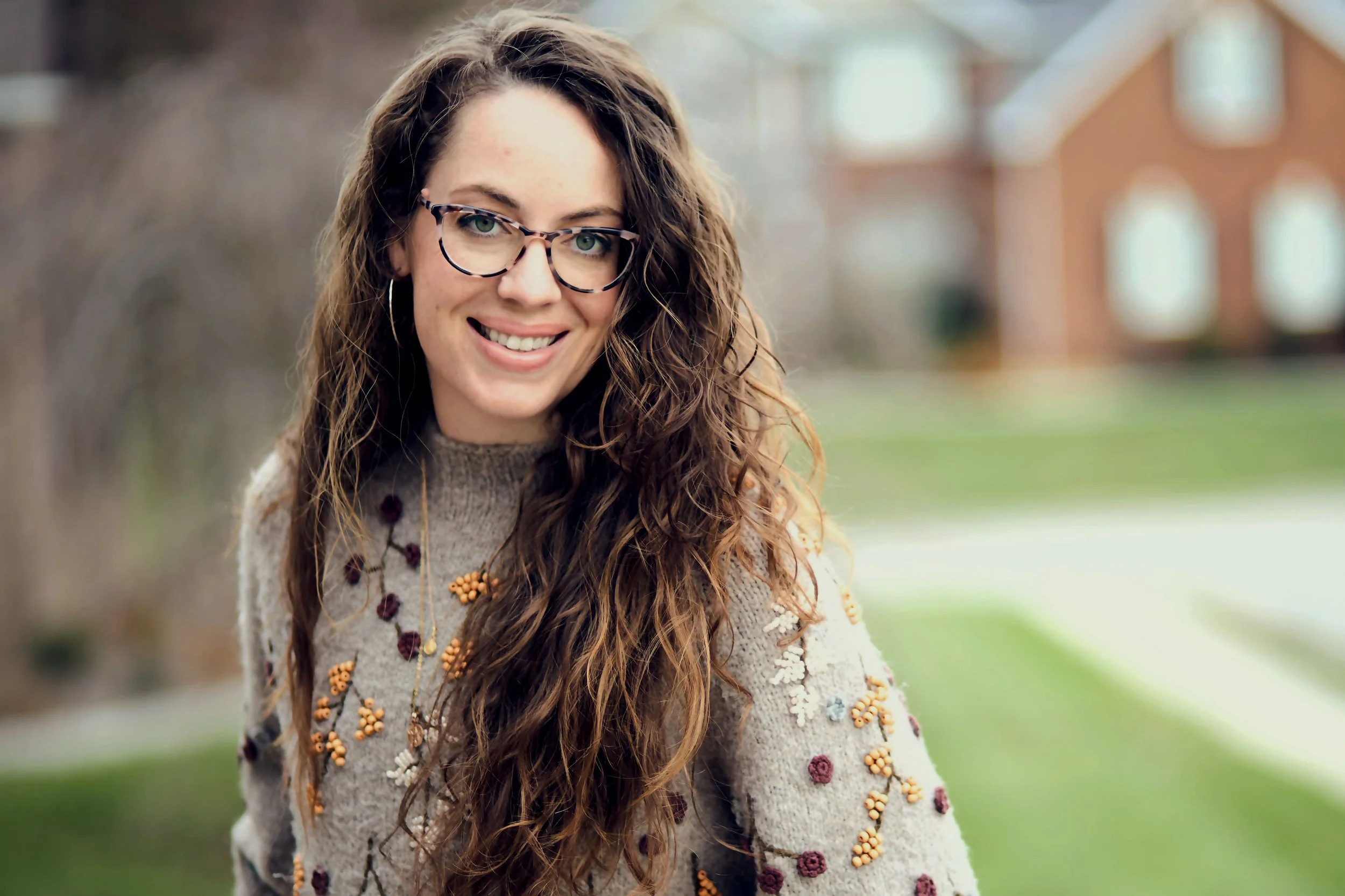 A smiling woman with long, curly brown hair and glasses outdoors during daytime, wearing a beige sweater with floral embroidery in a park setting.