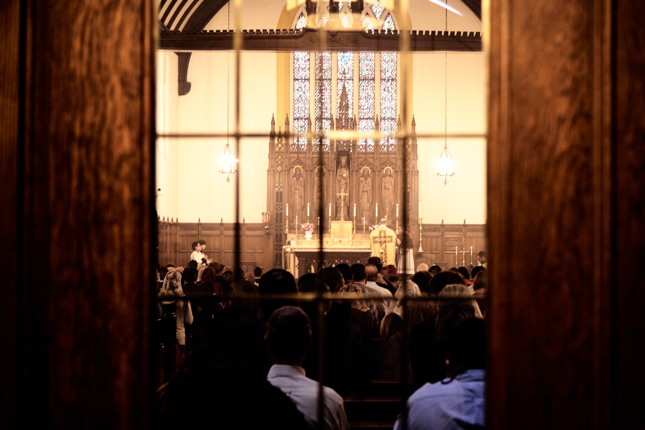 People gathered inside a church for a service, viewed through a window with a wooden frame. The church interior features stained glass windows and religious statues.