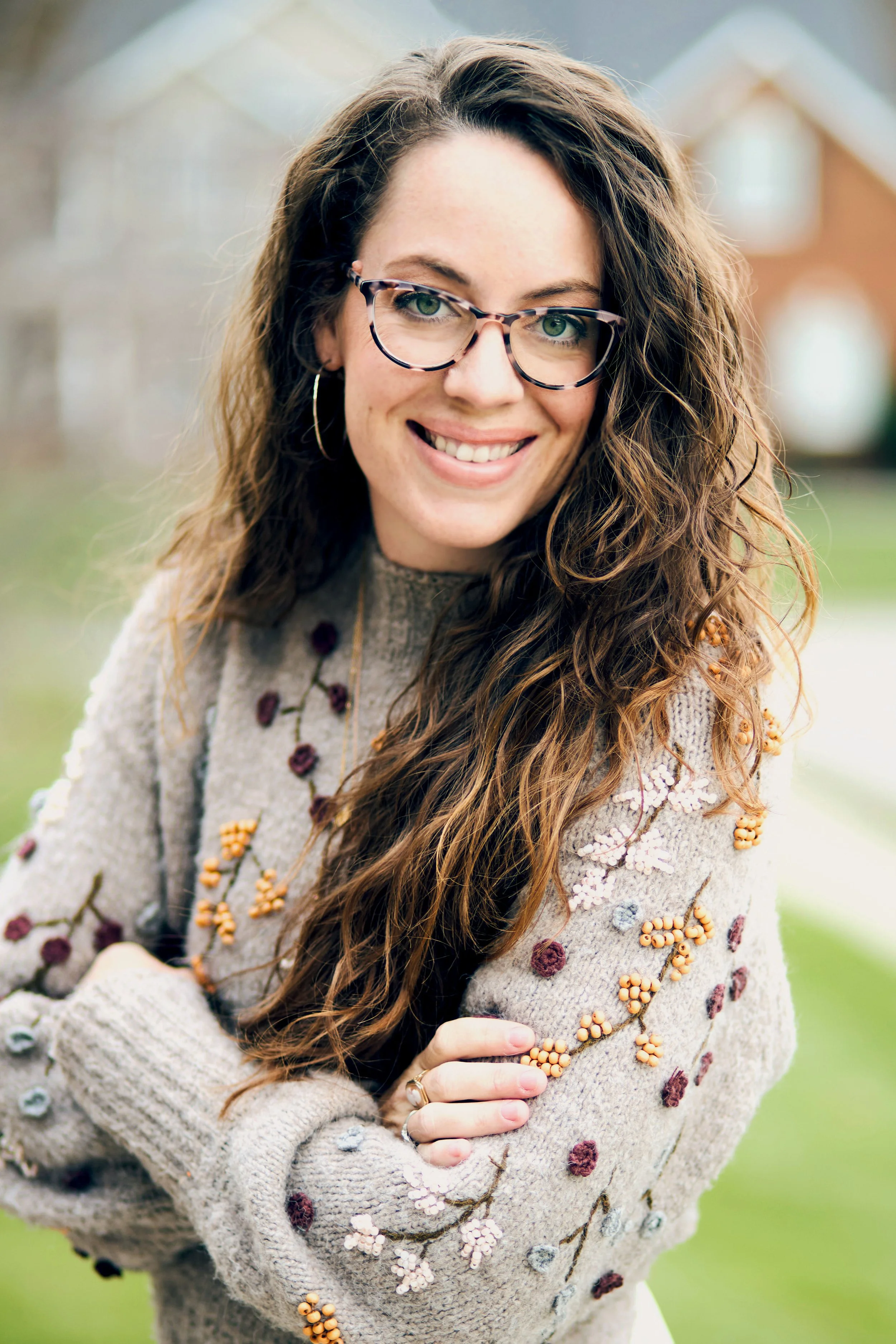 A woman with glasses and long curly brown hair smiling outdoors, wearing a beige sweater embroidered with colorful flowers and berries.