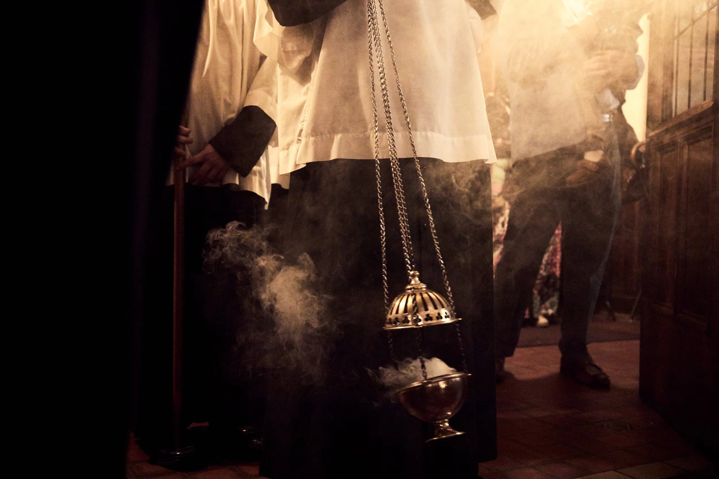 A person standing indoors wearing a long cream-colored coat with black cuffs, a chain strap bag hanging from the shoulder, and dark pants. The scene appears to be in a dimly lit room with wooden flooring and a vintage-style hanging lamp emitting warm light.