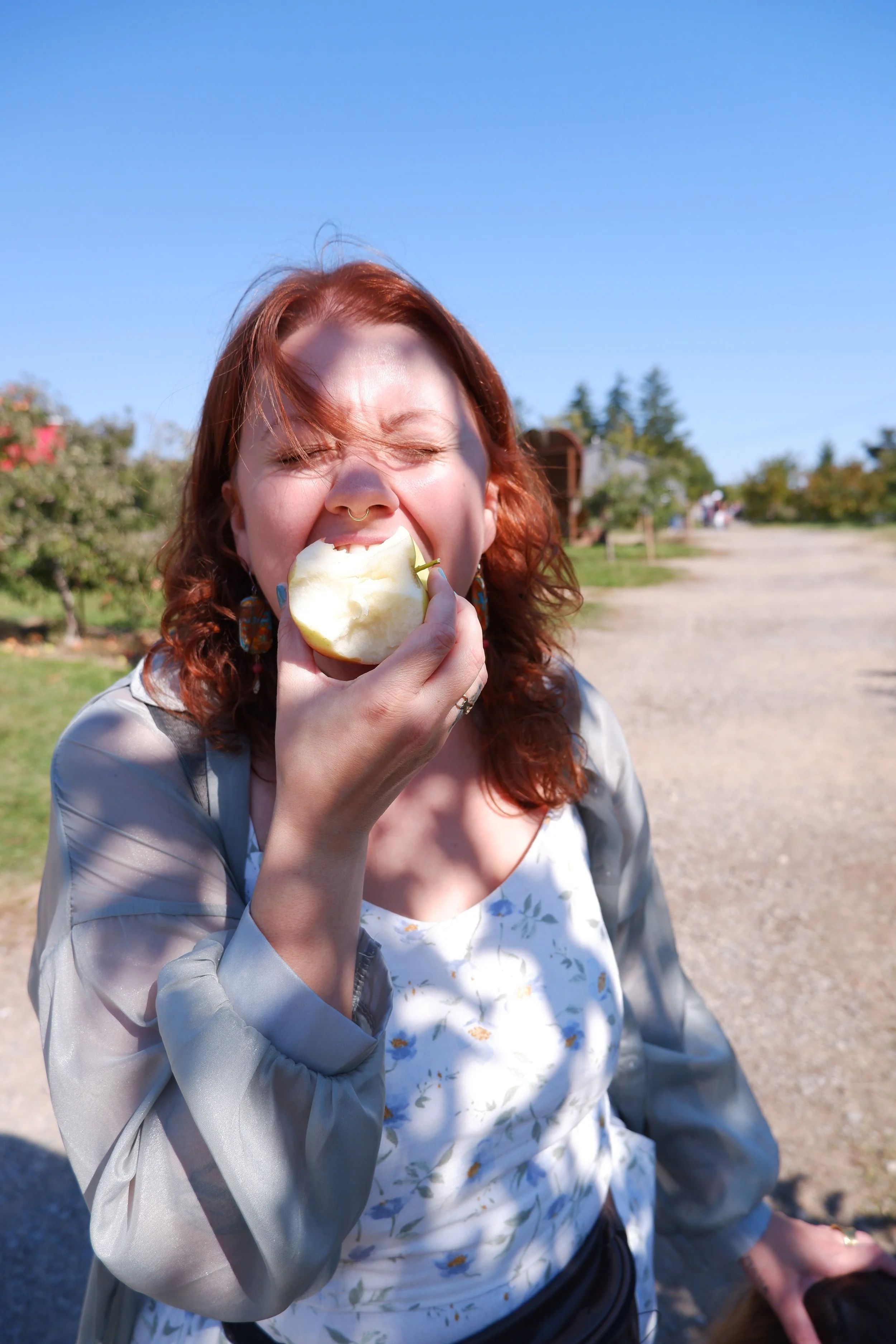 Woman with red hair and a septum piercing eating a green apple outdoors on a sunny day, with trees and a blue sky in the background.