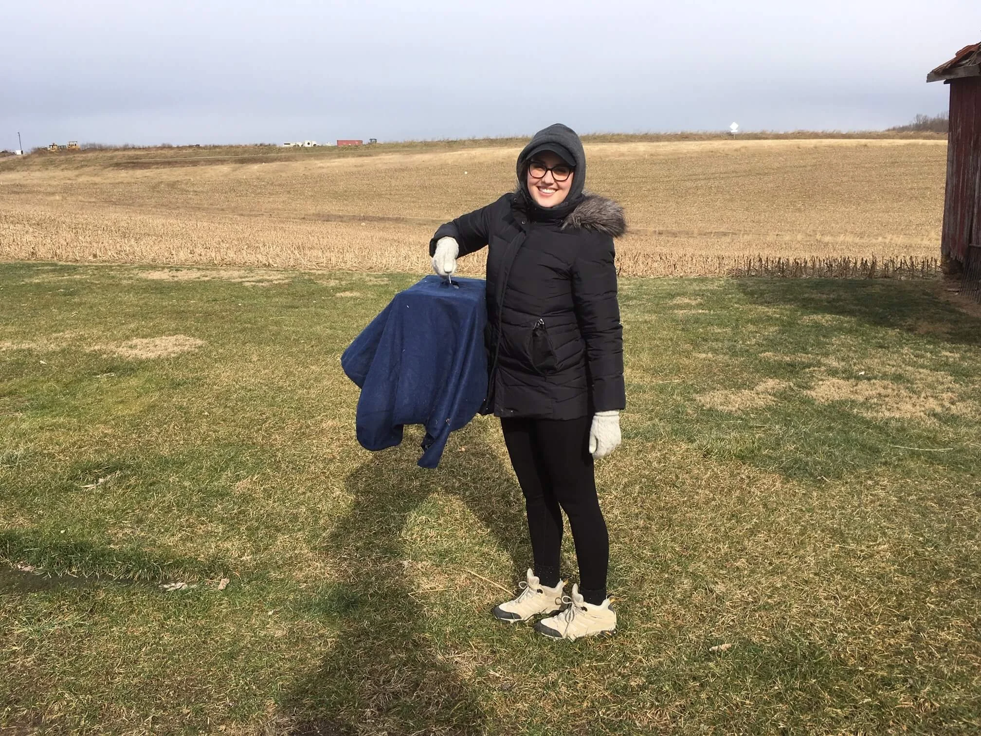 A woman in a black winter coat, gray hoodie, glasses, and white sneakers stands outdoors on a grassy area, smiling. She is holding a large blue bag. The background features a field with brown crops, a cloudy sky, and a red barn on the right side.