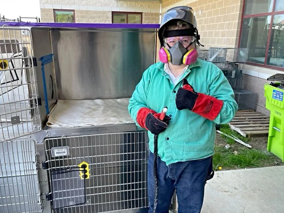 Person wearing a protective face mask, goggles, and gloves, standing next to a metal cage or kennel outdoors.