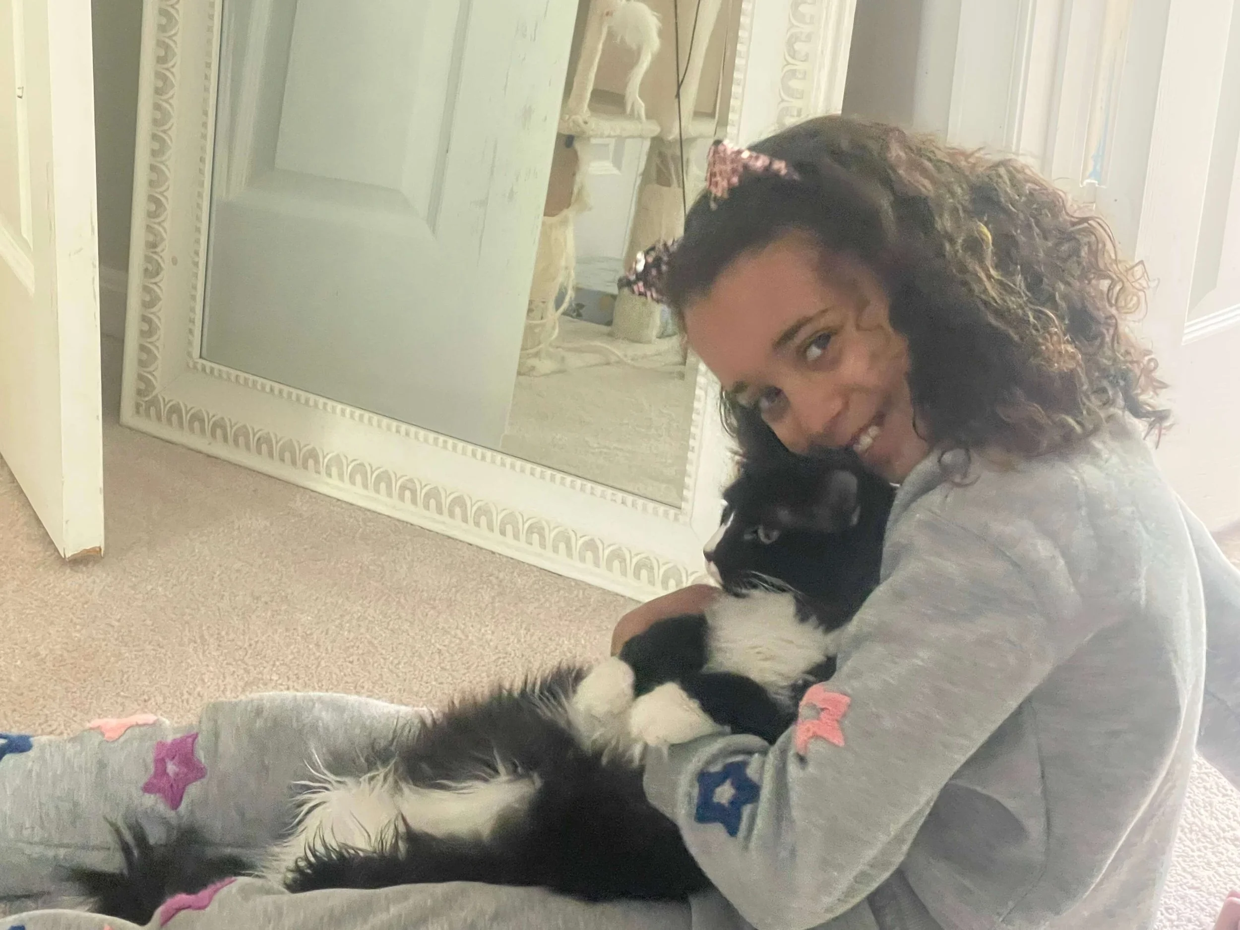 A woman with curly hair and a hair bow smiling and holding a black and white puppy inside a house near a large decorative mirror.