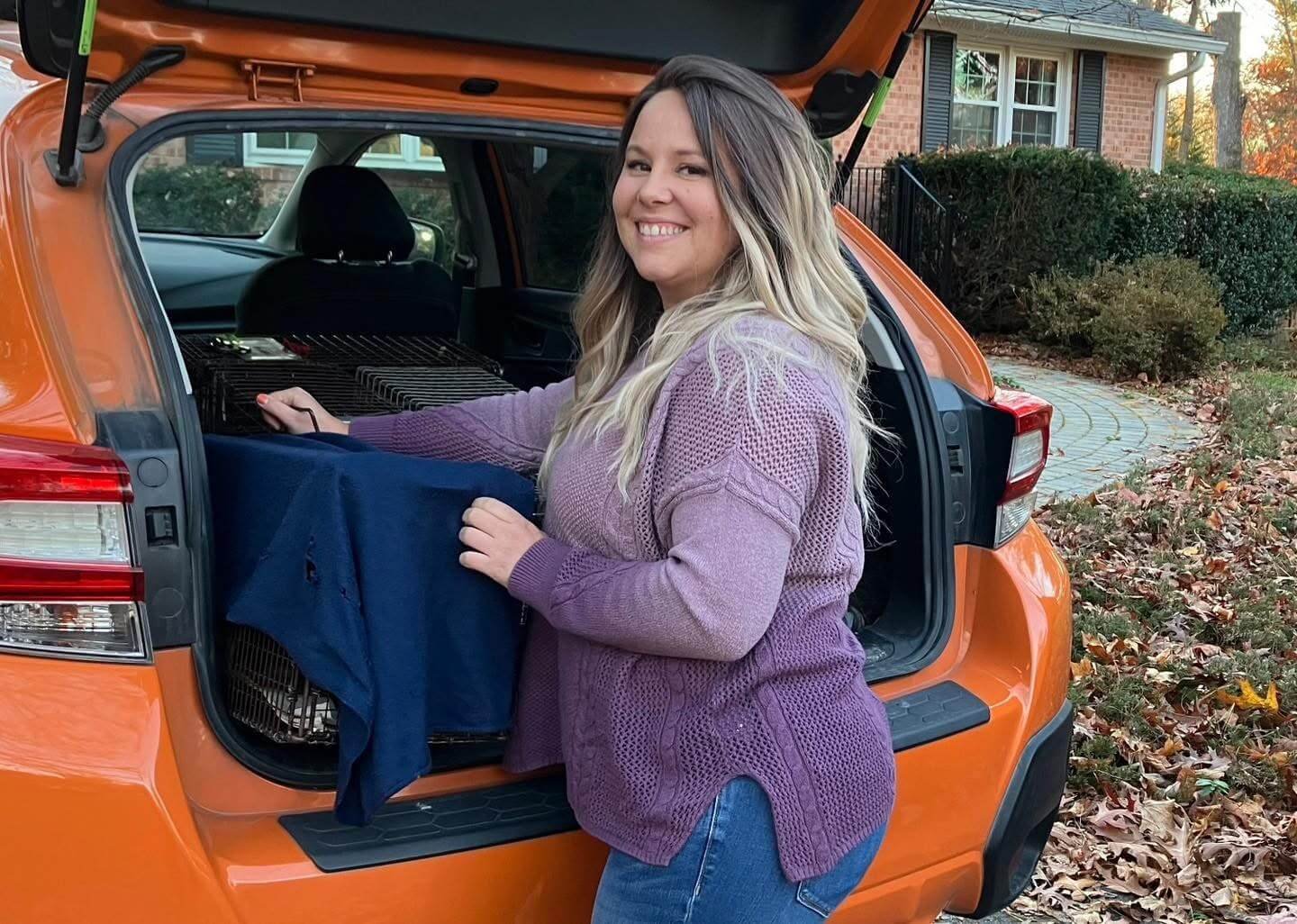 A woman loading a pet crate into the trunk of an orange SUV, parked outside in a yard with fallen autumn leaves and a brick house in the background.