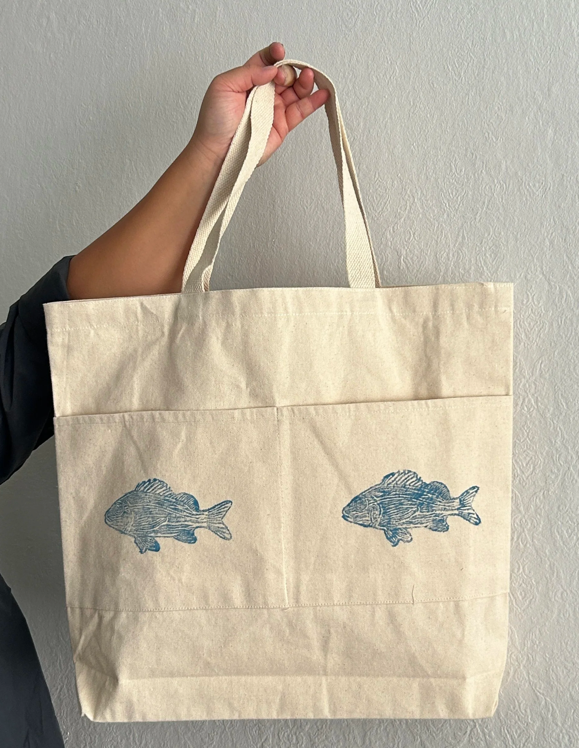 Person holding a beige tote bag with blue fish lino prints on a plain wall background.