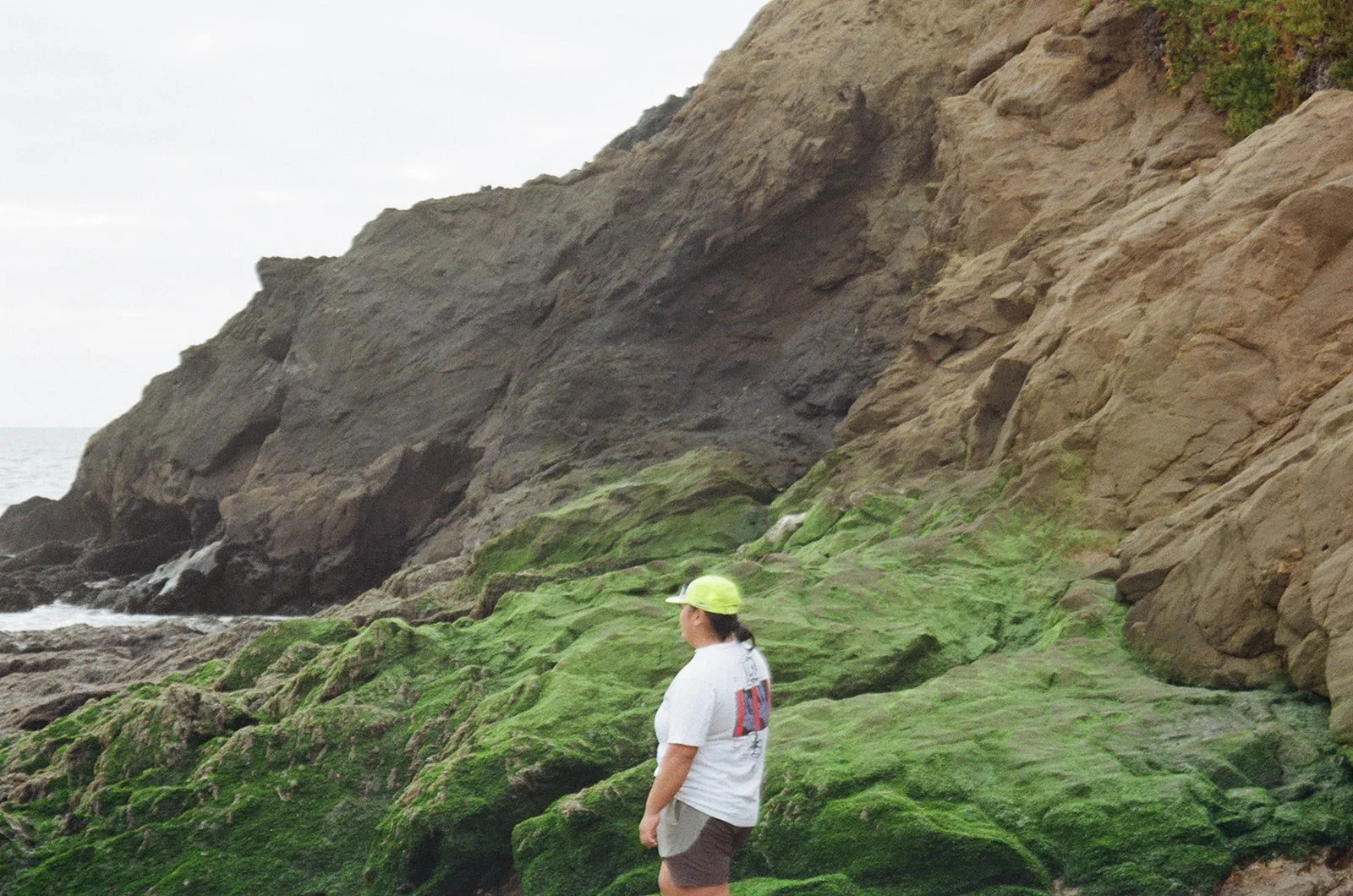 Person wearing a white T-shirt, gray shorts, and a yellow-green hat standing on moss-covered rocks near a rocky shoreline with ocean in the background.