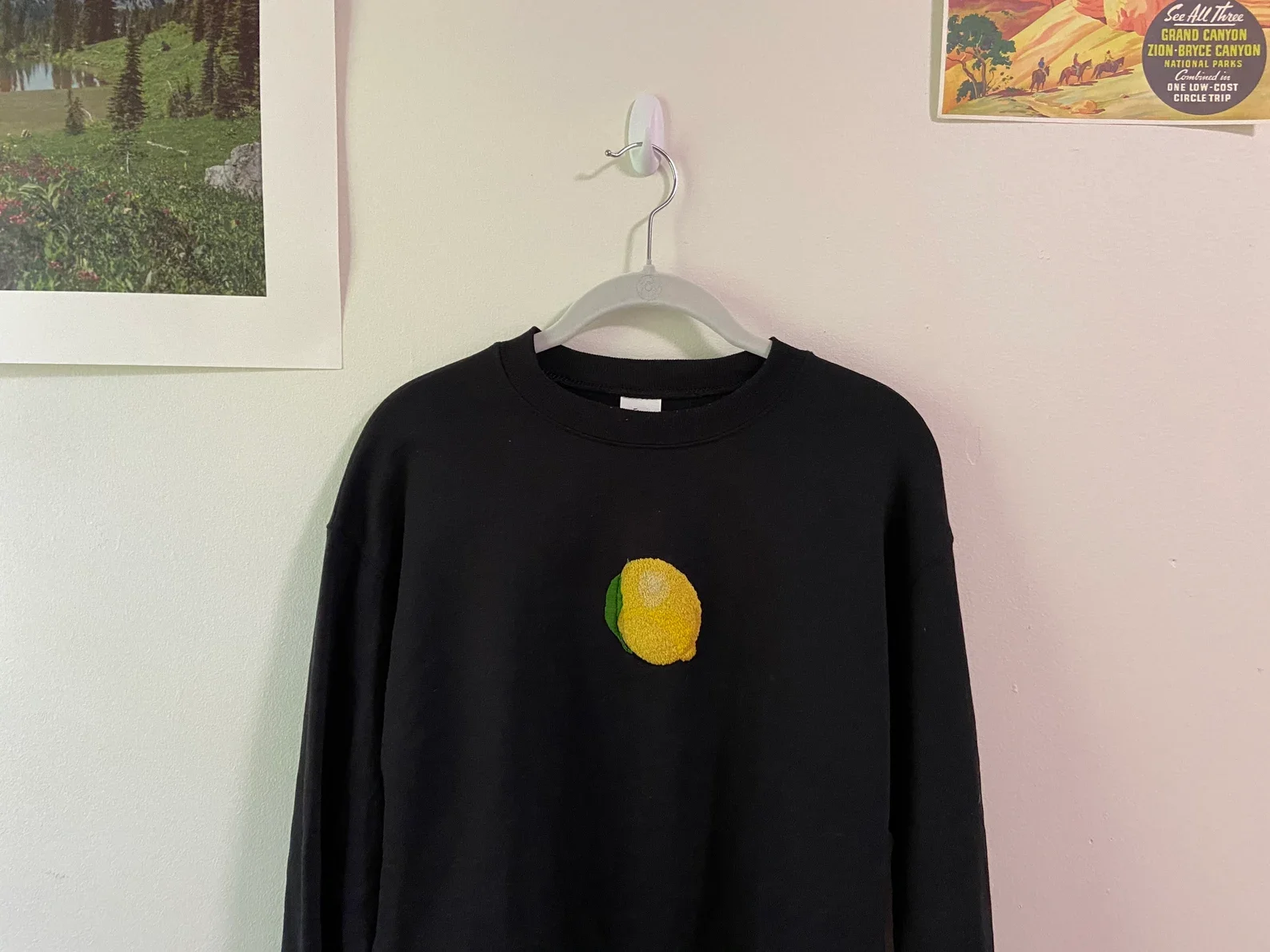 Black sweatshirt hanging on a white hanger on a wall. The sweatshirt has a yellow lemon embroidered on the chest.
