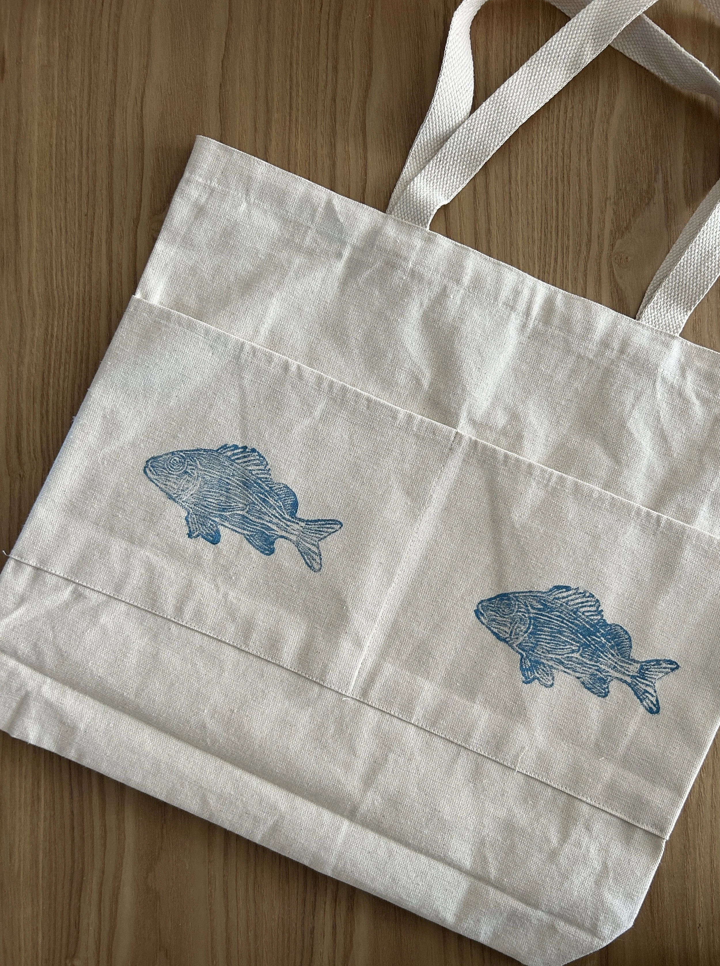 A white tote bag with two blue fish lino prints on the front, lying on a wooden surface.