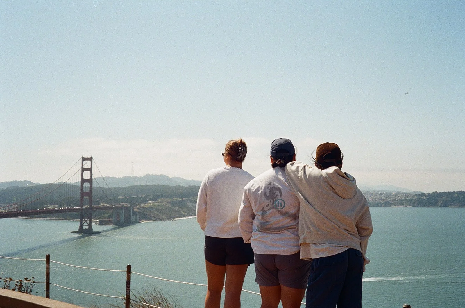 Three people stand side by side, looking at the Golden Gate Bridge over water on a clear day