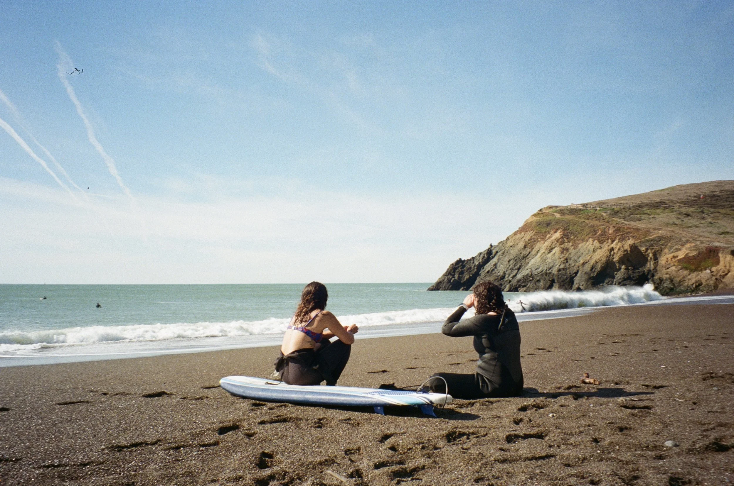 Two women sitting on a black sand beach near the shoreline, one with a surfboard, with the ocean, a rocky cliff, and a blue sky in the background.