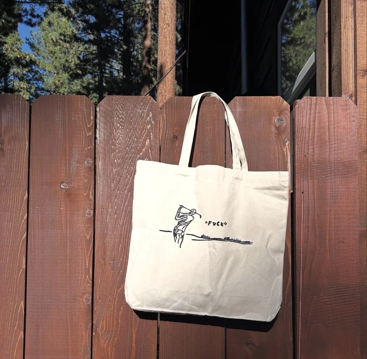 A white tote bag hanging on a wooden fence with a drawing of a person swinging a golf club and the word 'FUCK' written beside.