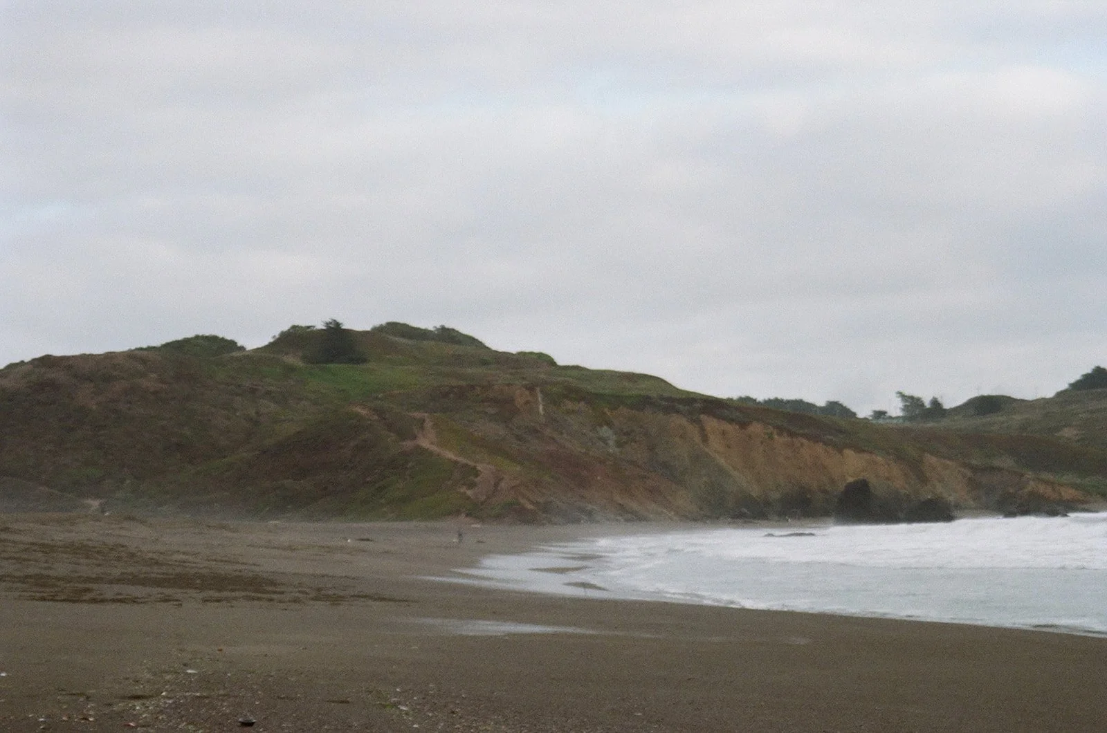 Beach with dark sand and waves, green hills in the background under a cloudy sky.
