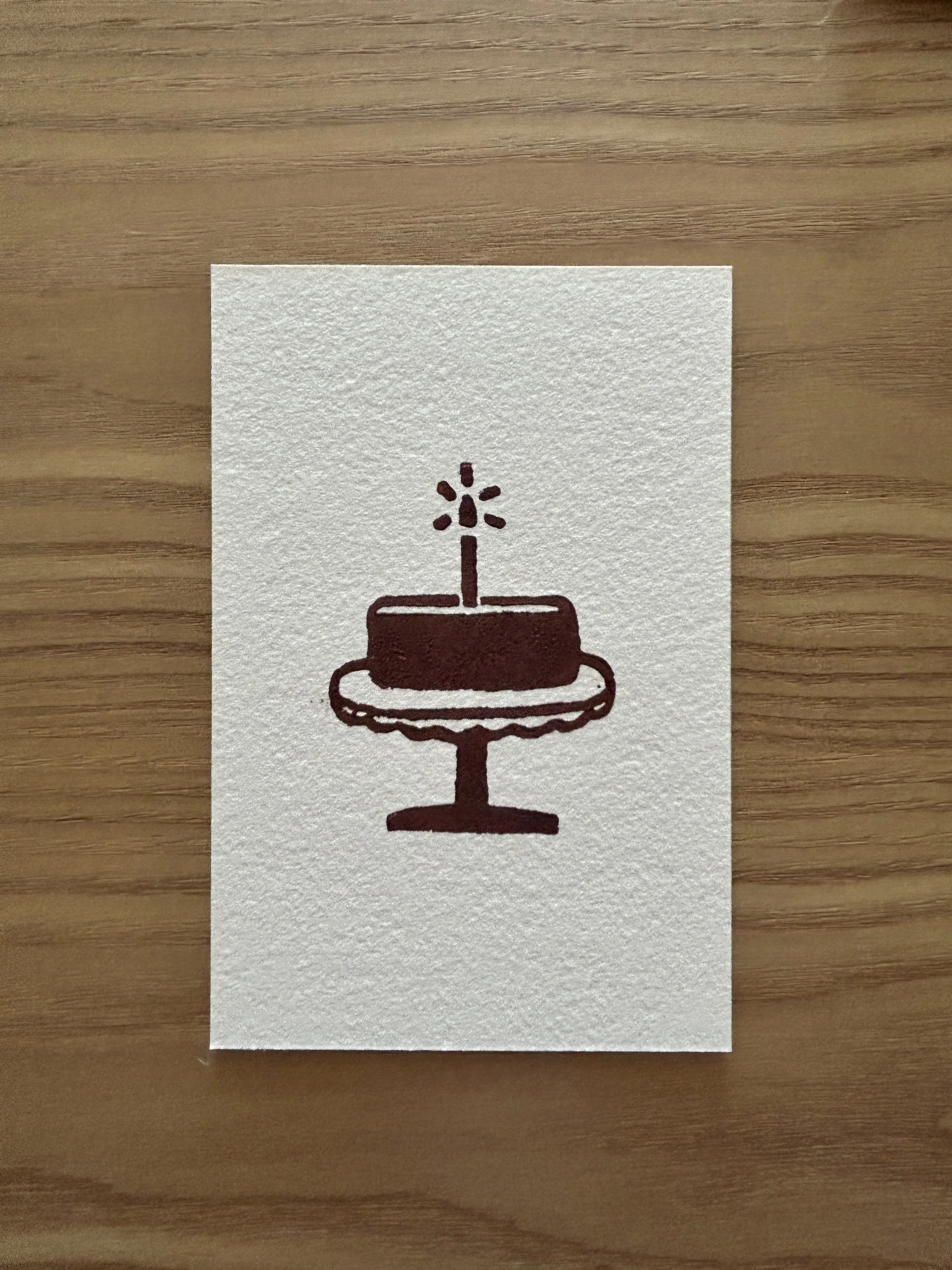 Lino printed birthday cake with a single lit candle on top, displayed on a cake stand.