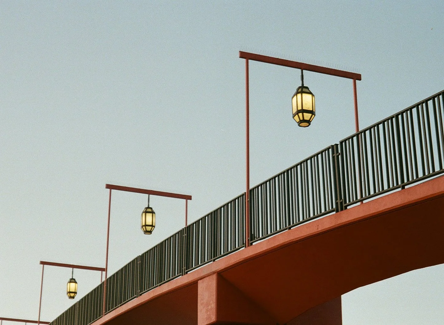 Photo of a bridge with black railings and red support beams, featuring hanging lantern-style lights against a light sky.