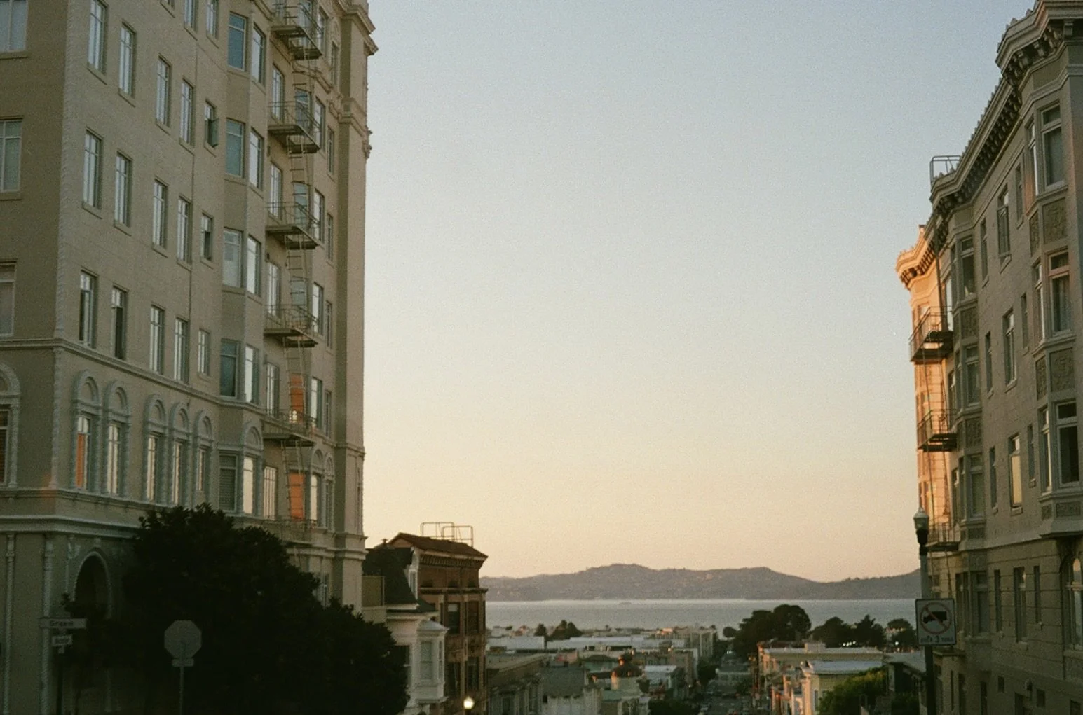 City street view with historic buildings on either side, overlooking a body of water and hills in the distance at sunset.