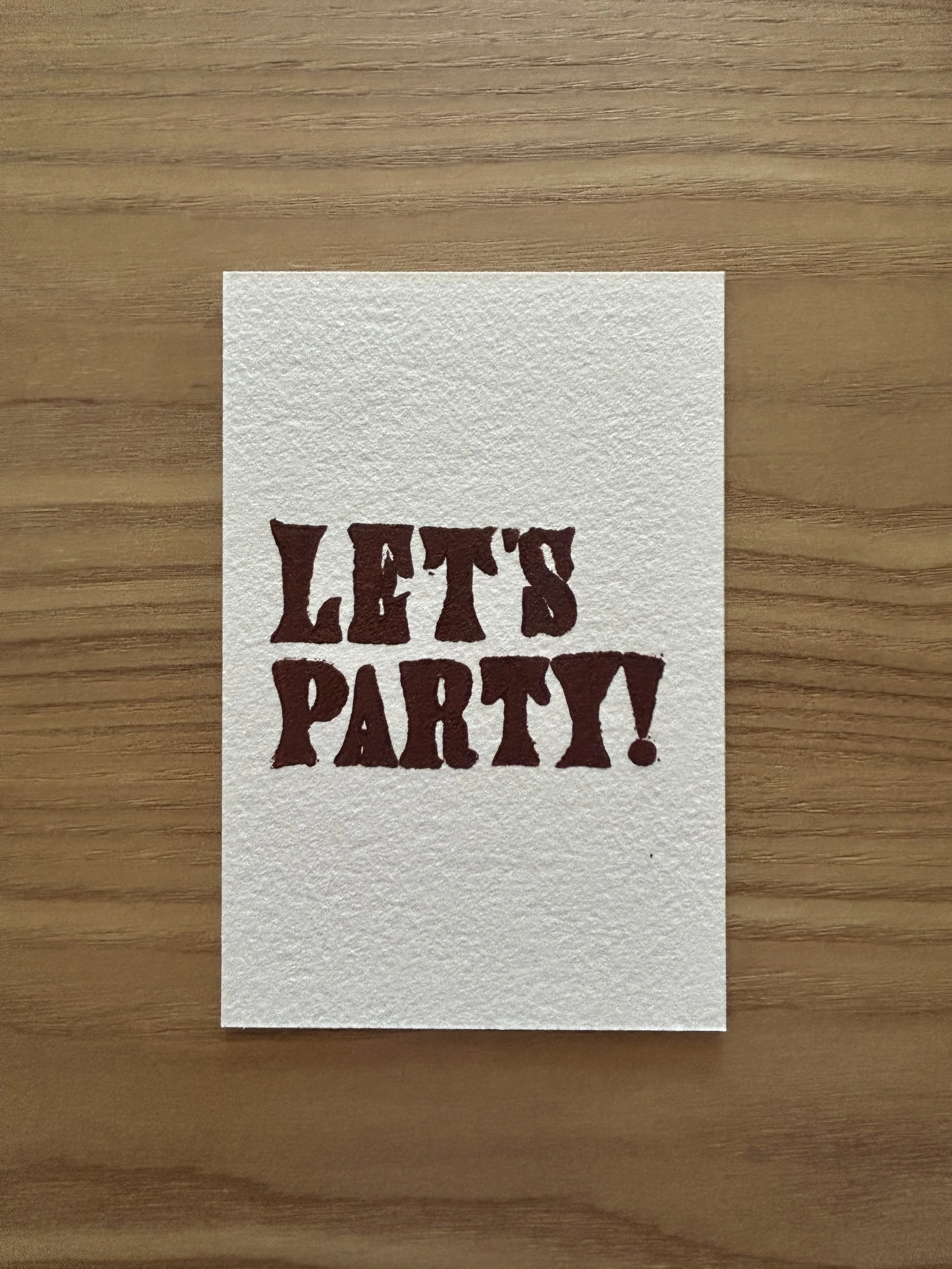 A white card with the words "LET'S PARTY!" printed in bold, dark red letters, placed on a wooden surface.