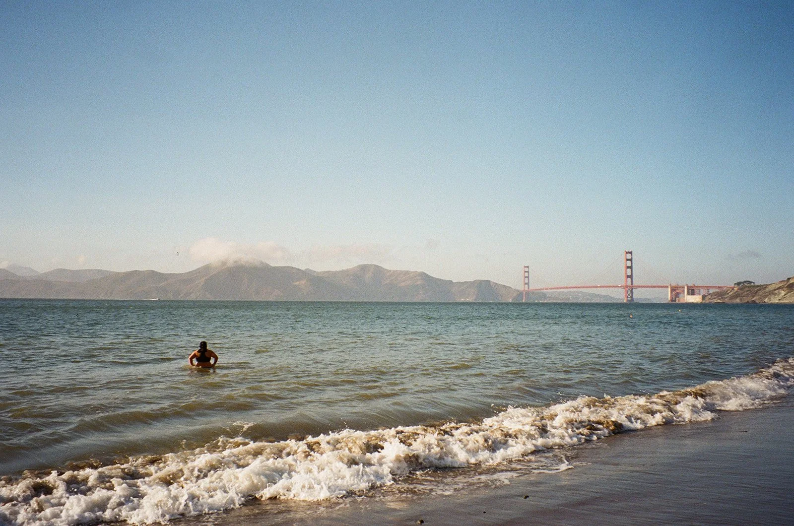 A woman swimming in the ocean near the shoreline with the Golden Gate Bridge visible in the background.