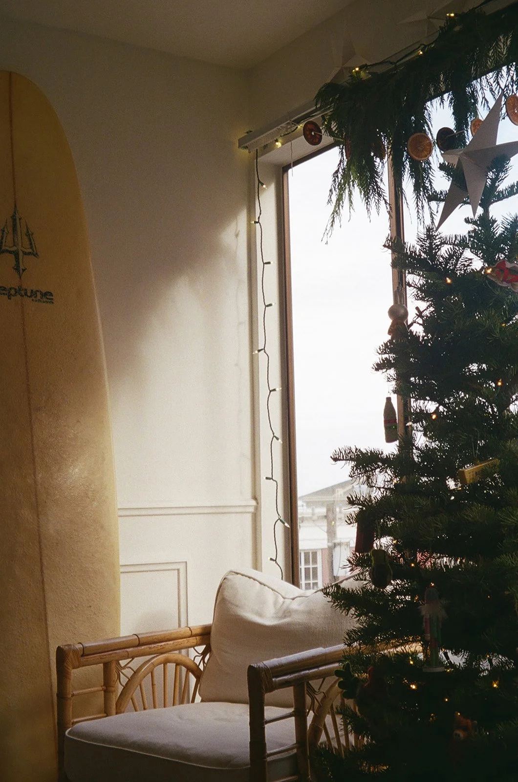 A cozy indoor scene with a Christmas tree decorated with ornaments and lights, near a window with a view of buildings outside. A wicker chair with white cushions is positioned next to the tree, and there are string lights hanging from the ceiling.