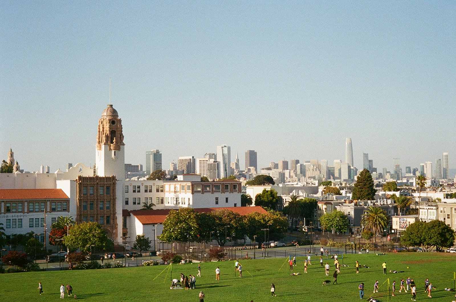 City skyline with a historic white building and a green park in the foreground, people playing sports on the grass.