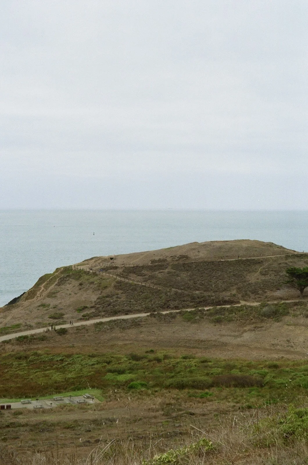Scenic view of a hillside near the ocean with walking trails and sparse vegetation under a cloudy sky.