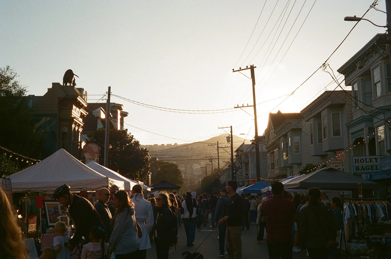 People walking and shopping at an outdoor street market during sunset, with tents, storefronts, and a cityscape in the background.