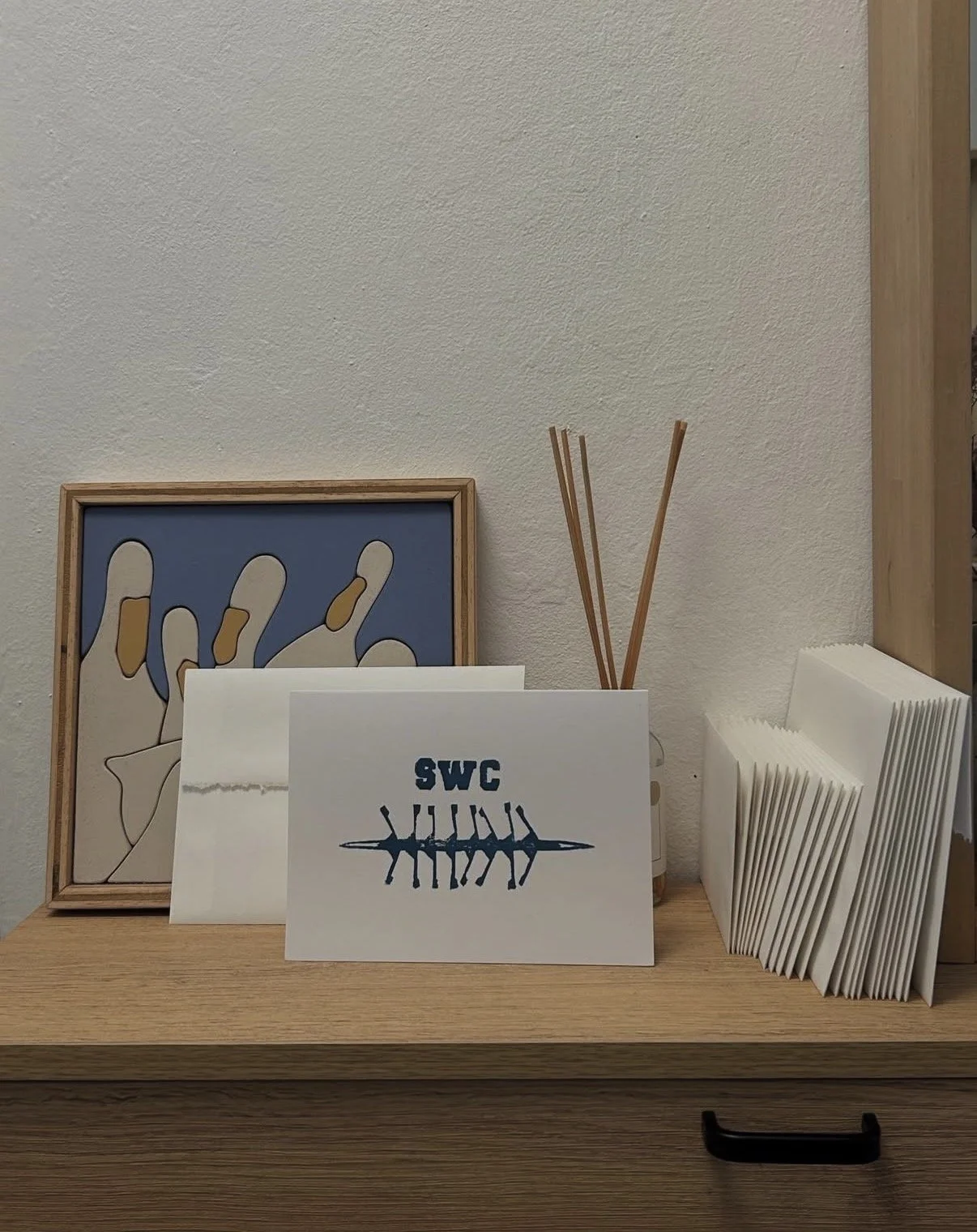 Wooden shelf with a framed abstract artwork, a white card with 'SWC' and a fishbone design, several blank white cards, and reed diffusers.