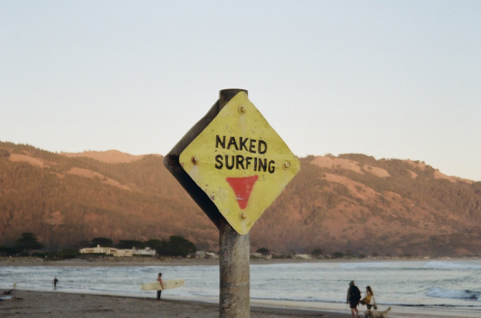 A yellow diamond-shaped sign that reads 'NAKED SURFING' with a red triangle below, located on a beach with surfers, people, and mountains in the background during sunset.