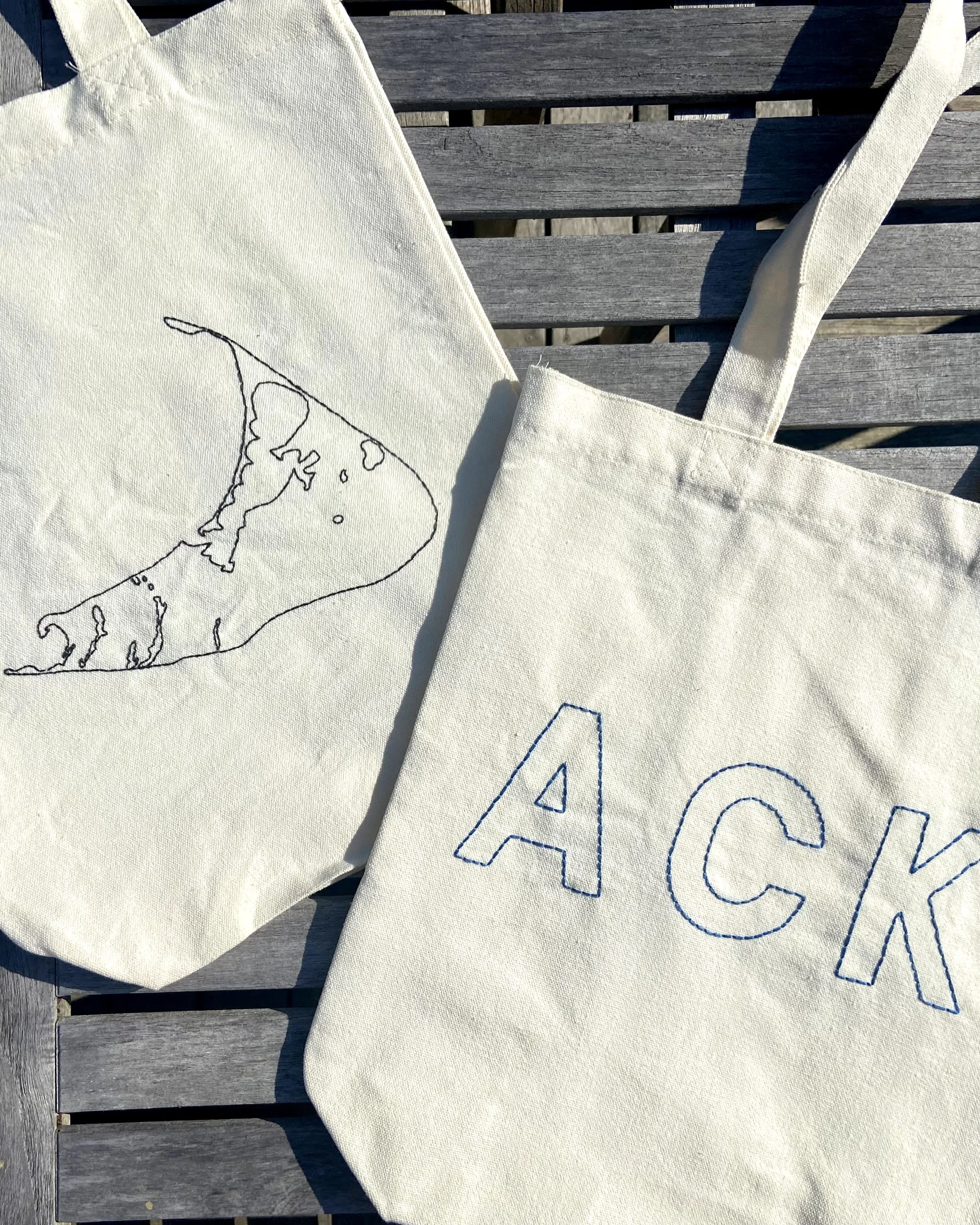 Two off-white fabric tote bags with embroidered designs resting on a wooden surface. One tote bag has the outline of Nantucket Island, and the other has the partially visible word "ACK" embroidered in blue.