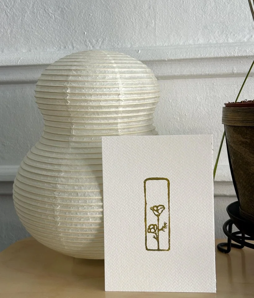 White paper lanterns, a small white card with a gold lino print of a flower, and a potted plant on a wooden surface.