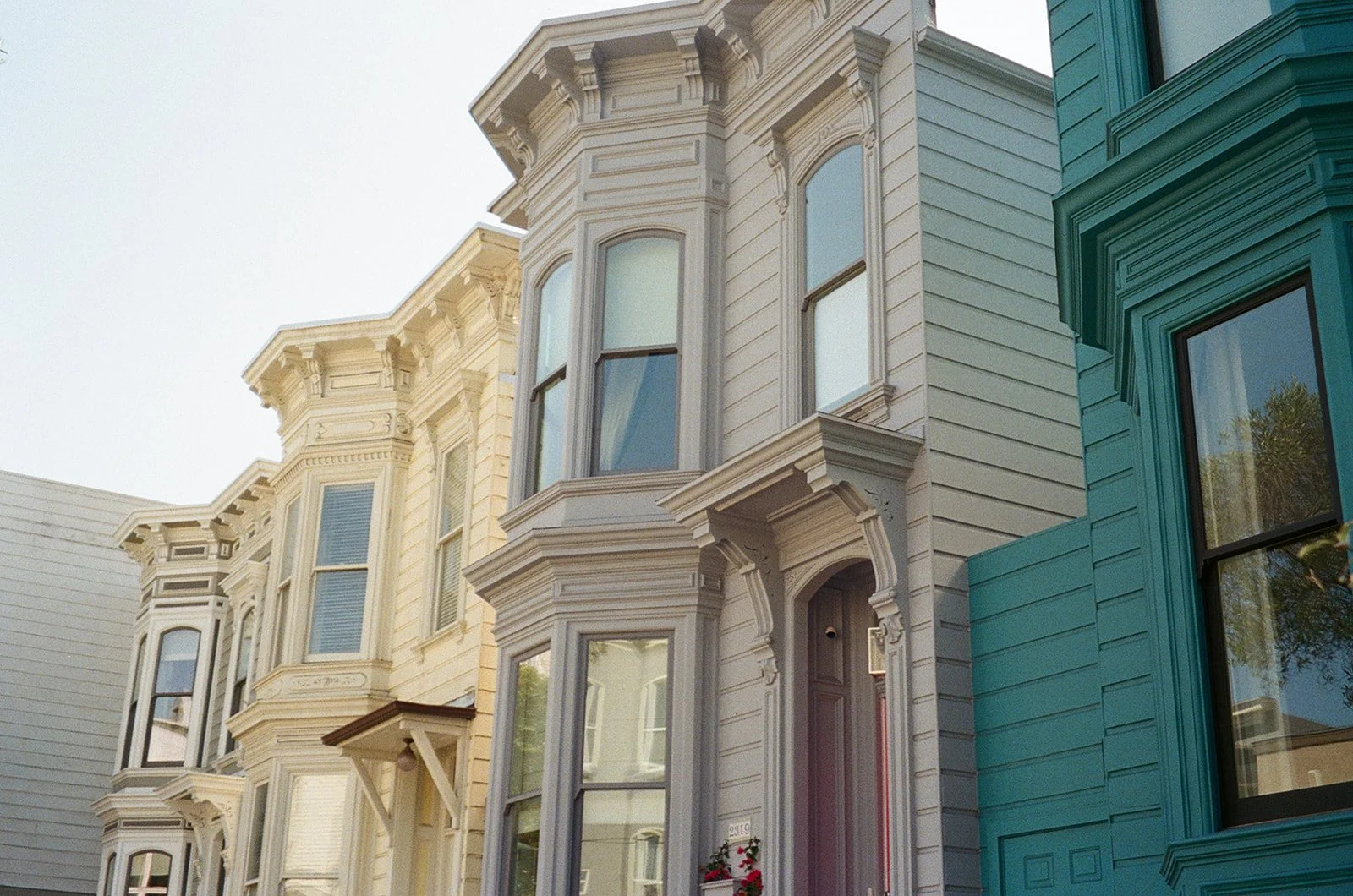 A row of Victorian-style houses with intricate trim and bay windows, painted in light and teal colors, on a sunny day.