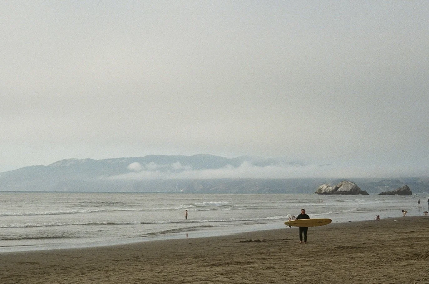 Person holding a surfboard walking on a sandy beach near the ocean with rocks and distant mountains in the background.