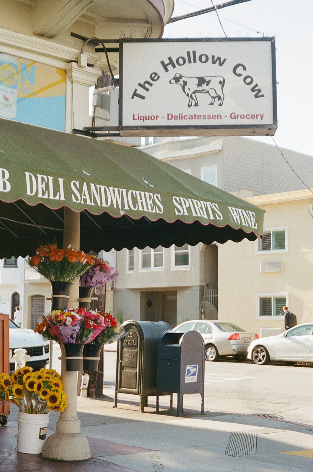Street view showing the sign for The Hollow Cow liquor, delicatessen, and grocery store, with fresh flowers displayed on the sidewalk and parked cars in the background.