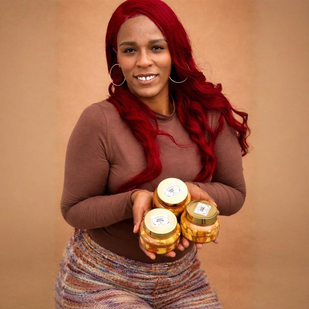 A woman with long red hair wearing hoop earrings, a brown long-sleeve shirt, and multicolored striped pants is holding four jars of food, smiling at the camera against a plain beige background.