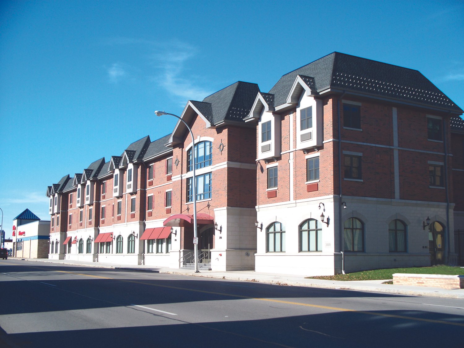 A multi-story brick building with arched windows and red awnings on the street under a clear blue sky.
