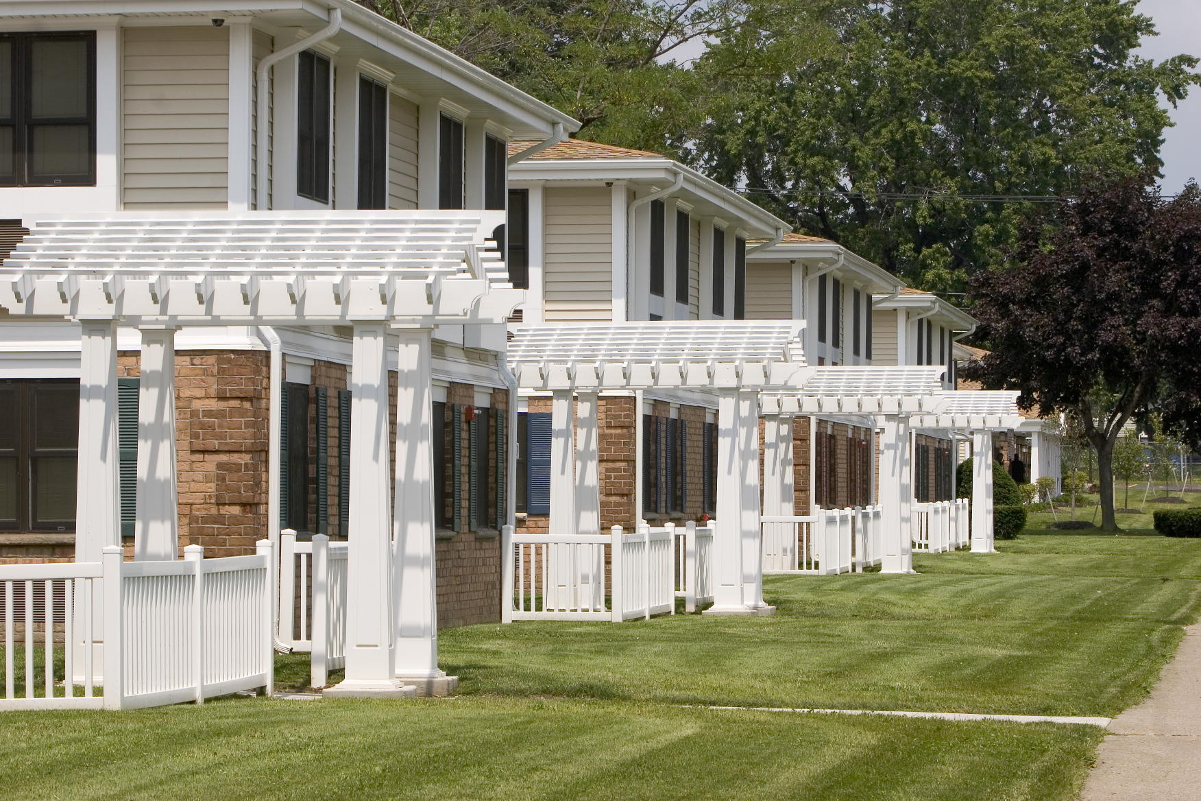 A row of modern townhouses with brick and siding exteriors, white pergolas on the porches, black window shutters, green lawns, and trees in the background.