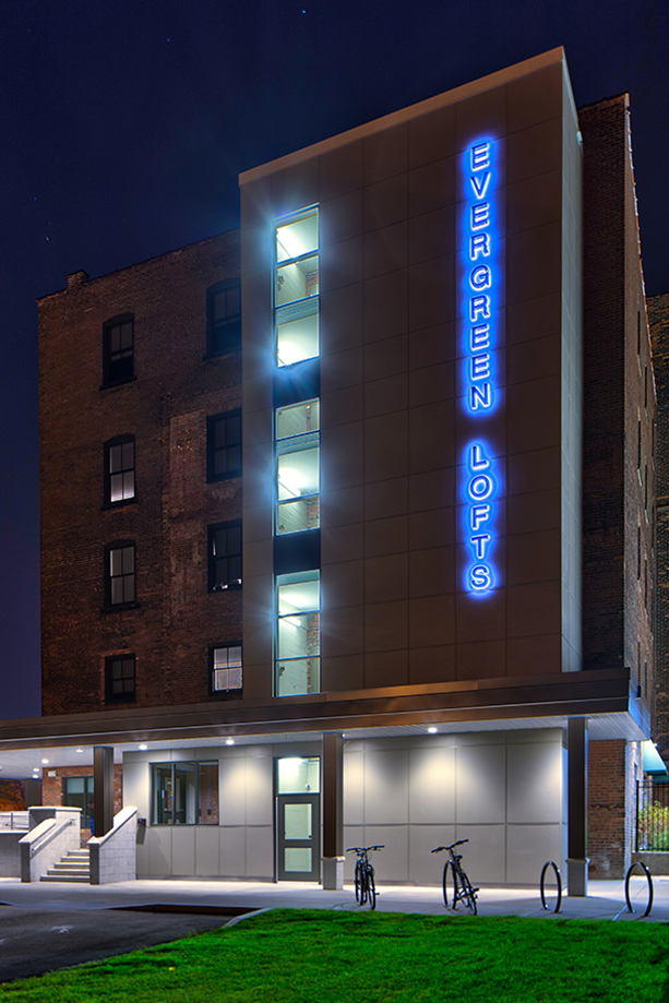 Night view of a modern building with a vertical blue neon sign reading "Evergreen Lofts," adjacent to an older brick building, with bicycles parked outside.