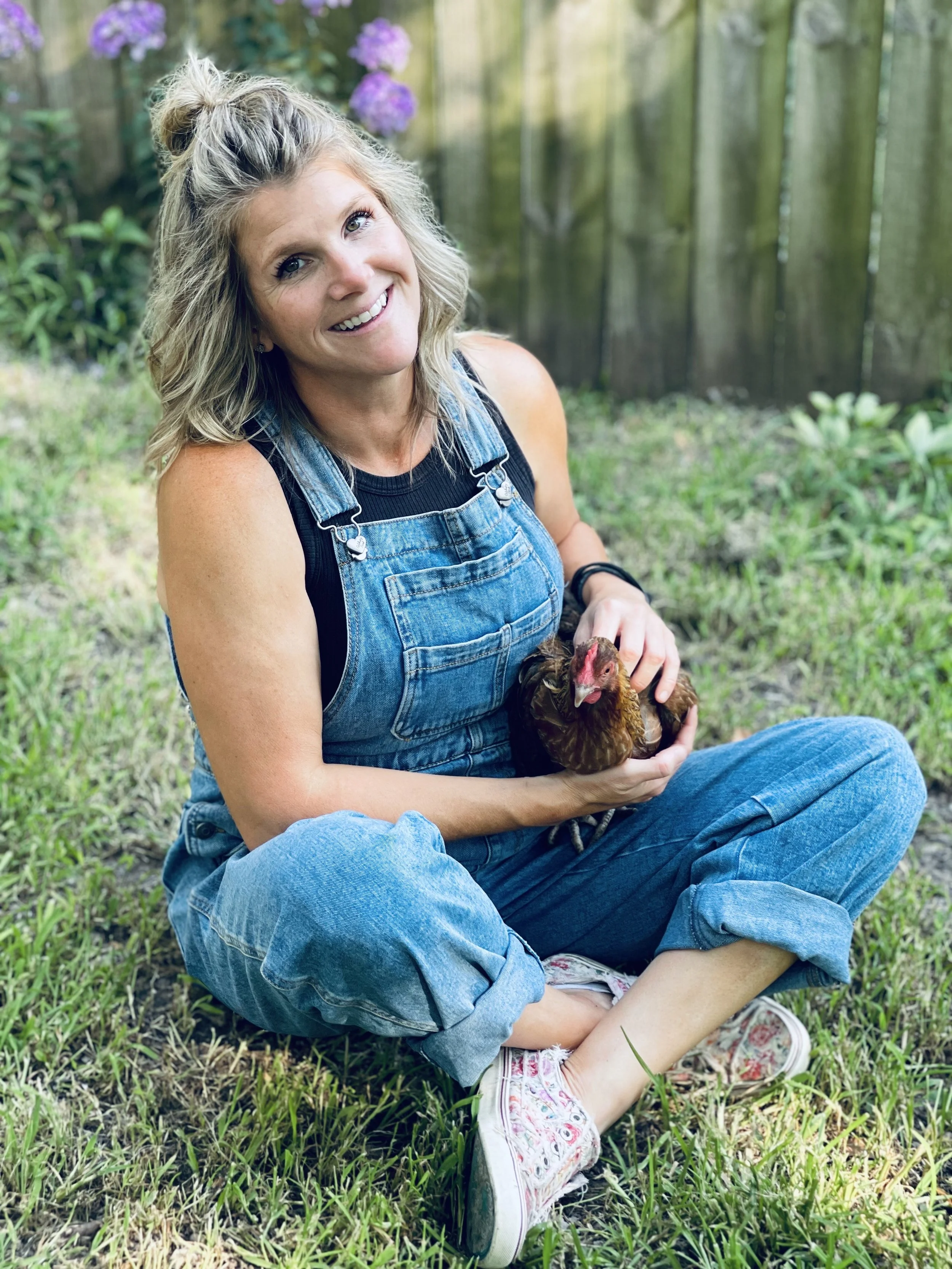 A woman with blonde hair in a messy bun, wearing denim overalls over a black top, sitting on the grass outdoors, holding a brown chicken in her hands, and smiling at the camera.