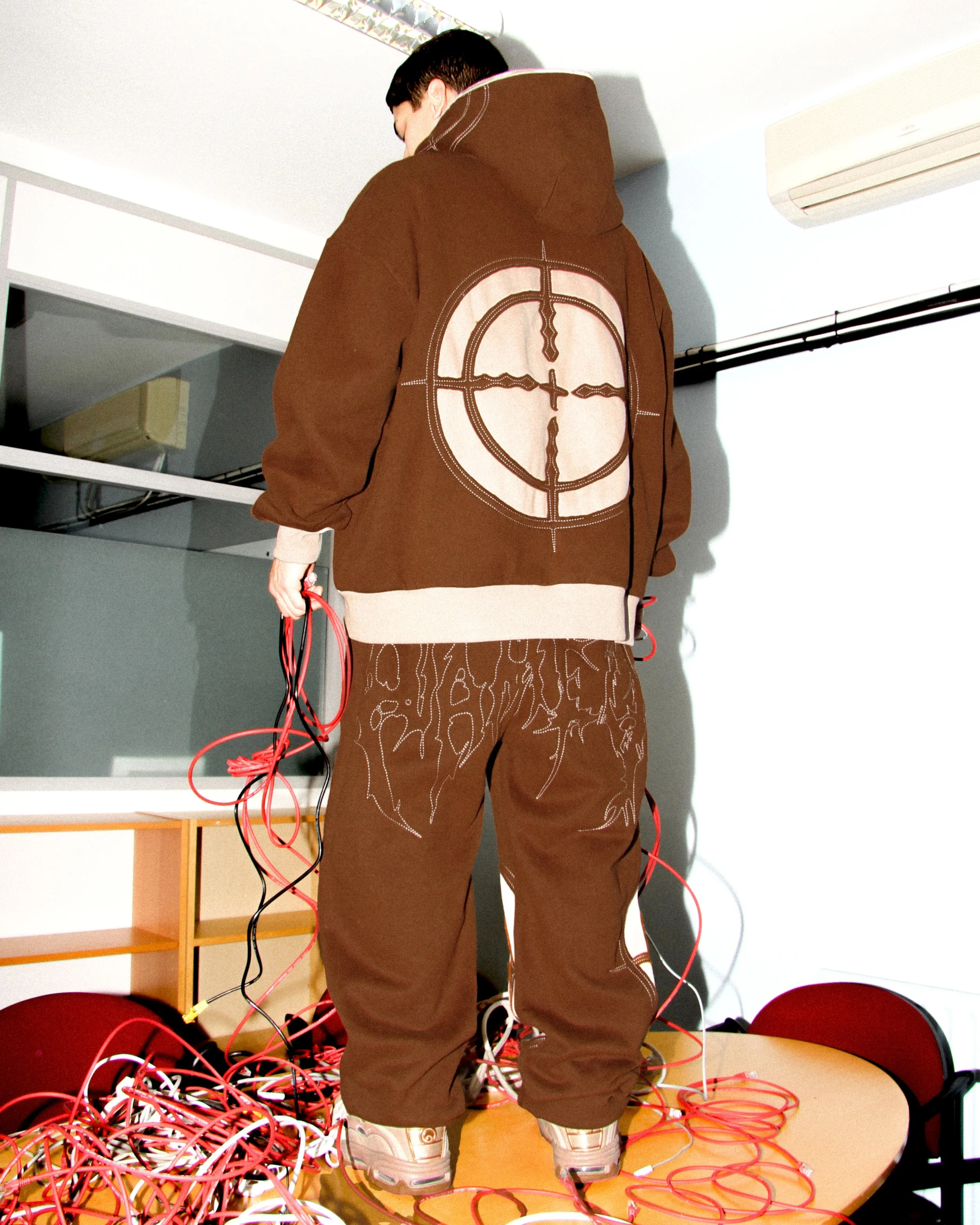 Launch campaign image for NAMED Collective “Classified” capturing the back graphic of a brown hoodie and joggers styled with tangled red cables in a tech-inspired studio environment.