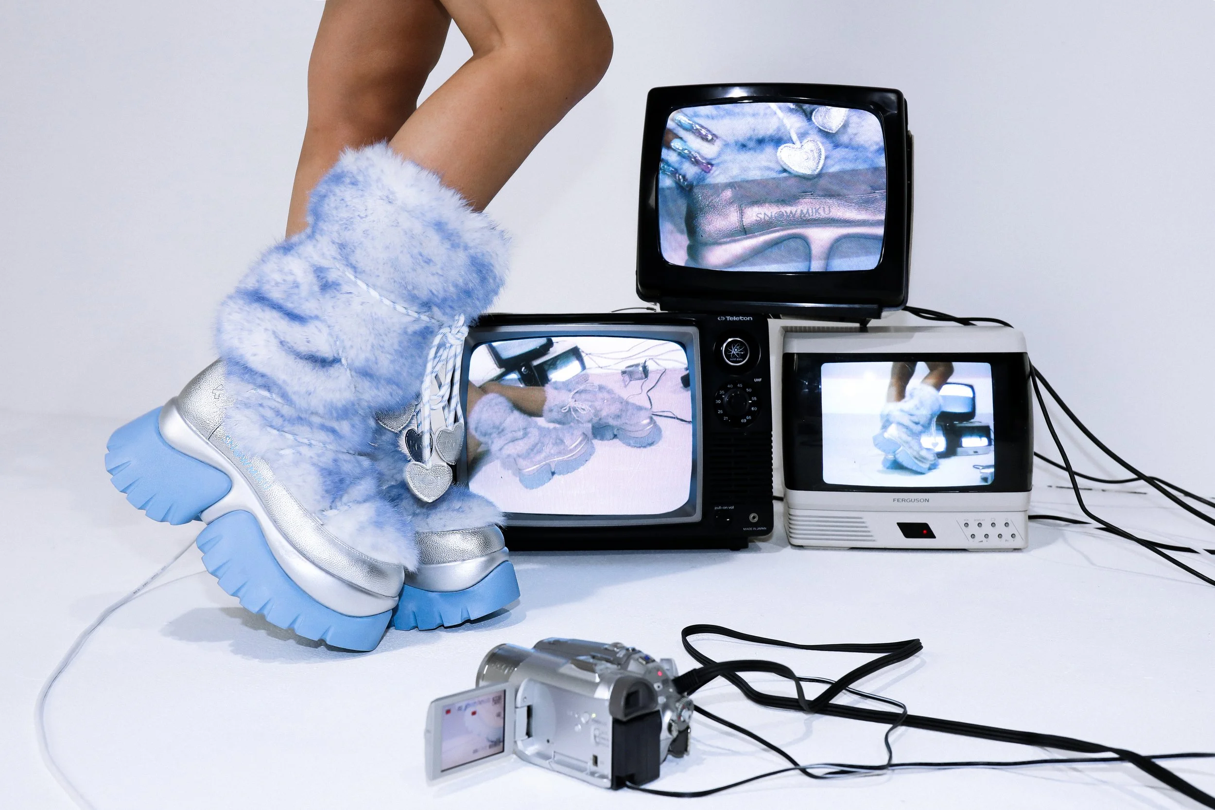 Hatsune Miku x KOI campaign imagery featuring faux-fur powder blue and metallic silver snow boots, styled within a technology-inspired white cove set surrounded by stacked CRT televisions and live VHS video feedback.