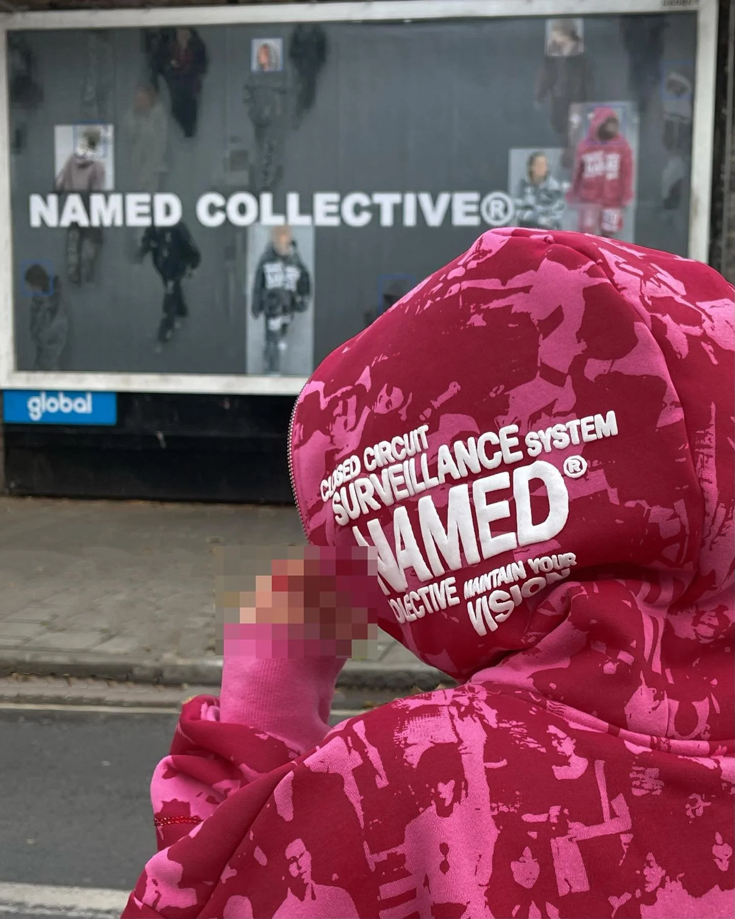Person wearing pink hoodie with white text, standing near bus stop with advertisement board featuring people and the text "NAMED COLLECTIVE".