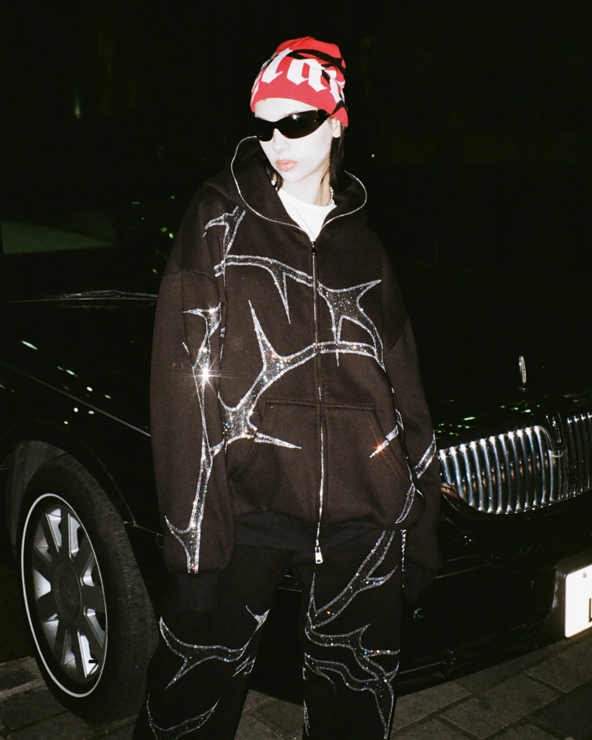 Female model poses in front of a black limousine in London for NAMED Collective's "Thorn" campaign, wearing a black rhinestone tracksuit, red and white logo beanie, and black sunglasses.