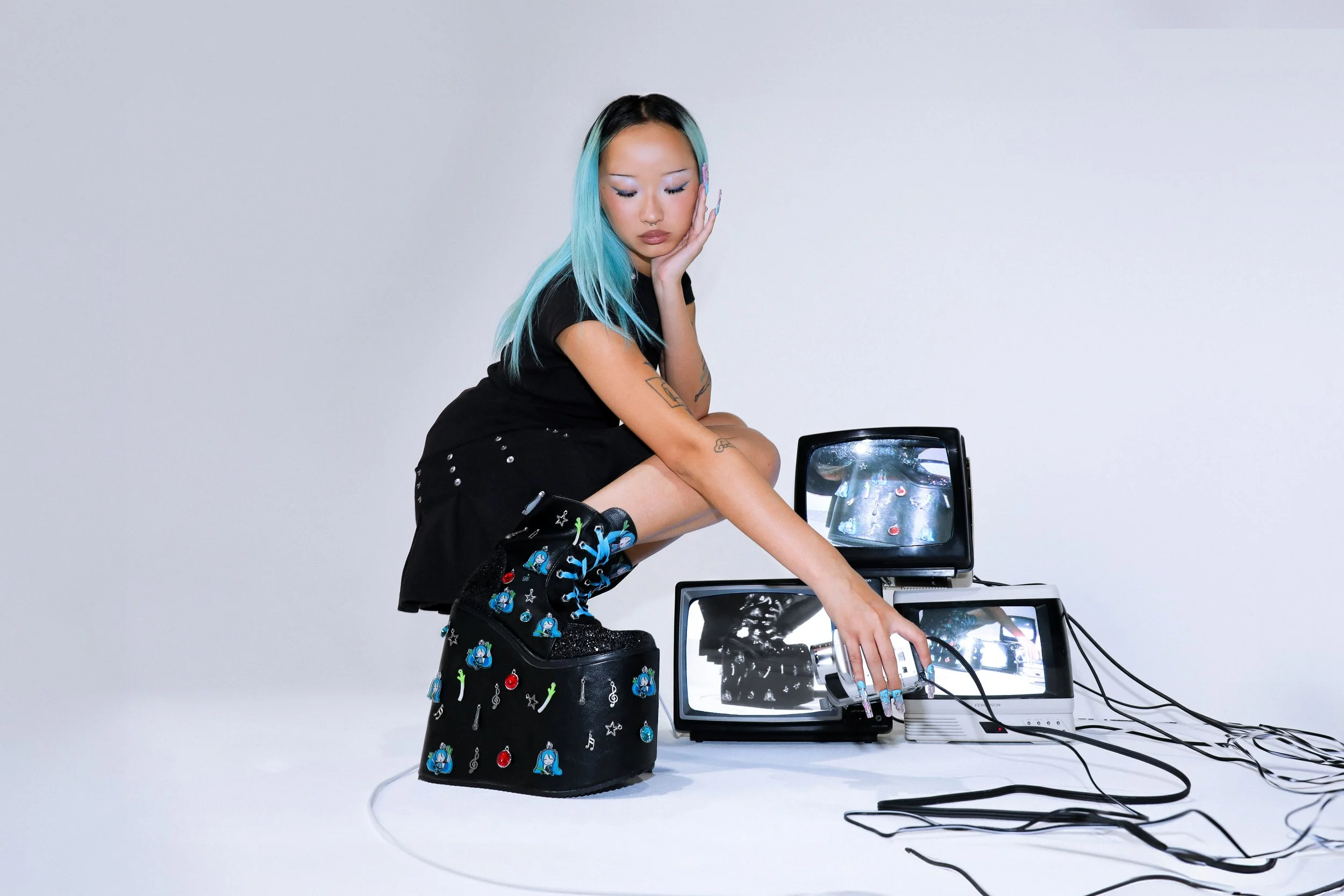 Hatsune Miku x KOI campaign imagery featuring a model wearing platform black boots adorned in charms, styled within a technology-inspired white cove set surrounded by stacked CRT televisions and live VHS video feedback.