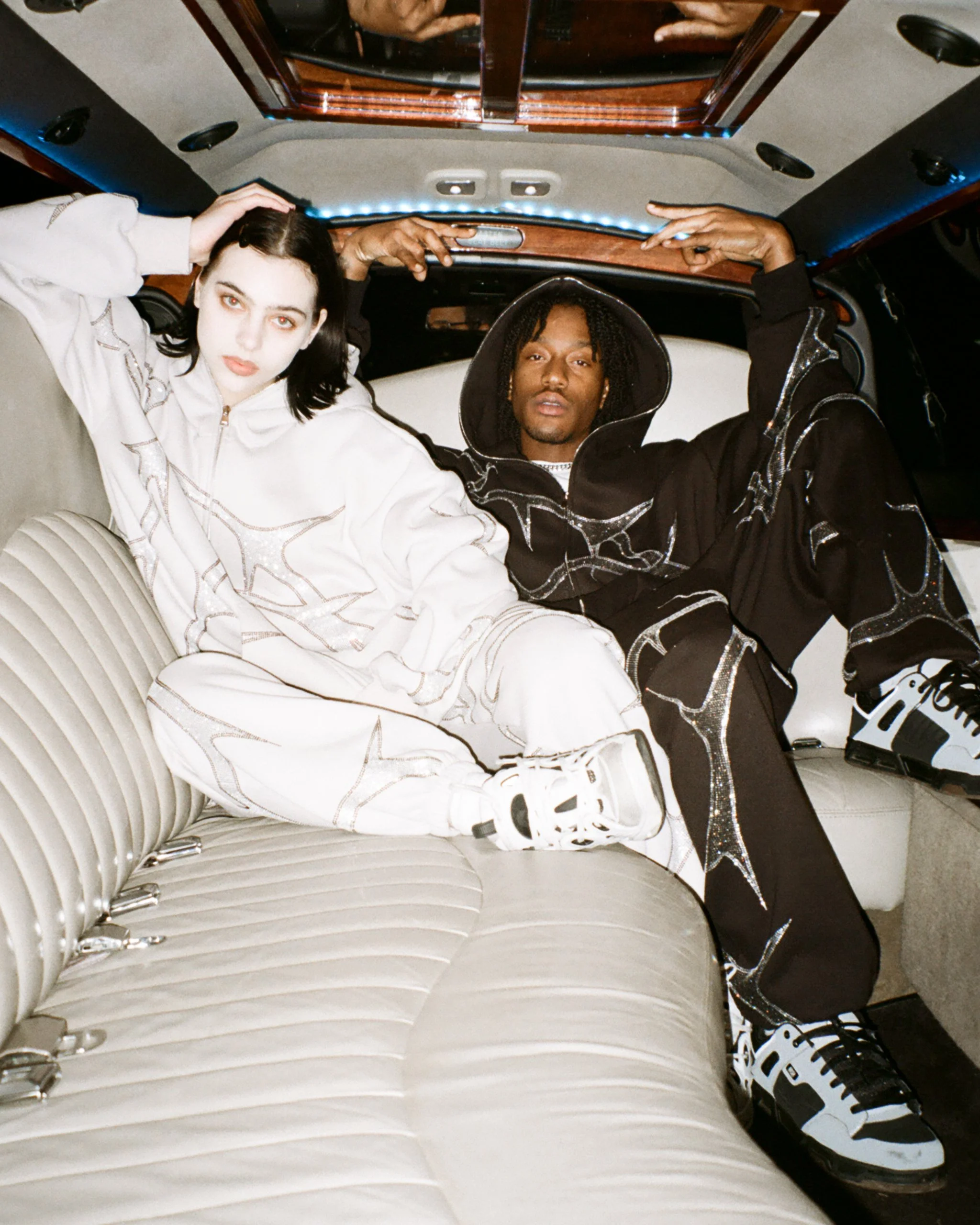 Female and male model posing inside a limousine with beige interiors, wearing a bone rhinestone tracksuit and black rhinestone tracksuit, for NAMED Collective's "Thorn" campaign.