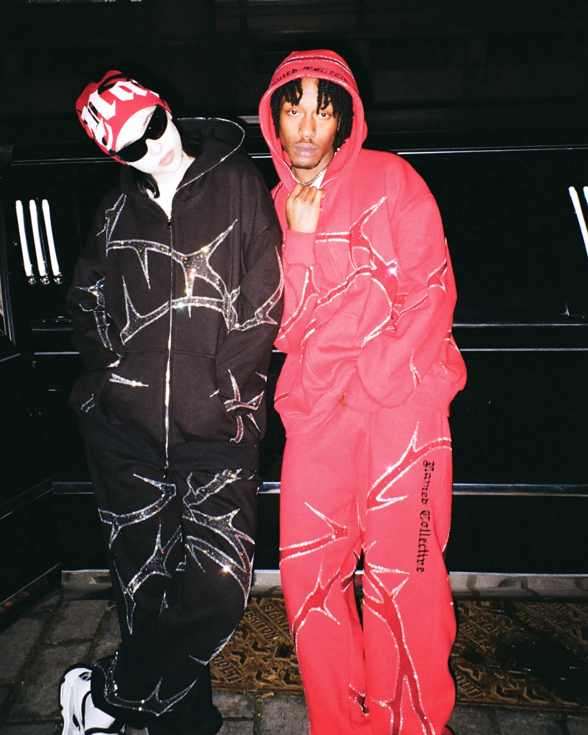 Female and male model posing in front of black limousine for NAMED Collective's "Thorn" campaign, wearing a black rhinestone tracksuit, red beanie hat and black sunglasses, and a red rhinestone tracksuit with the hood up.