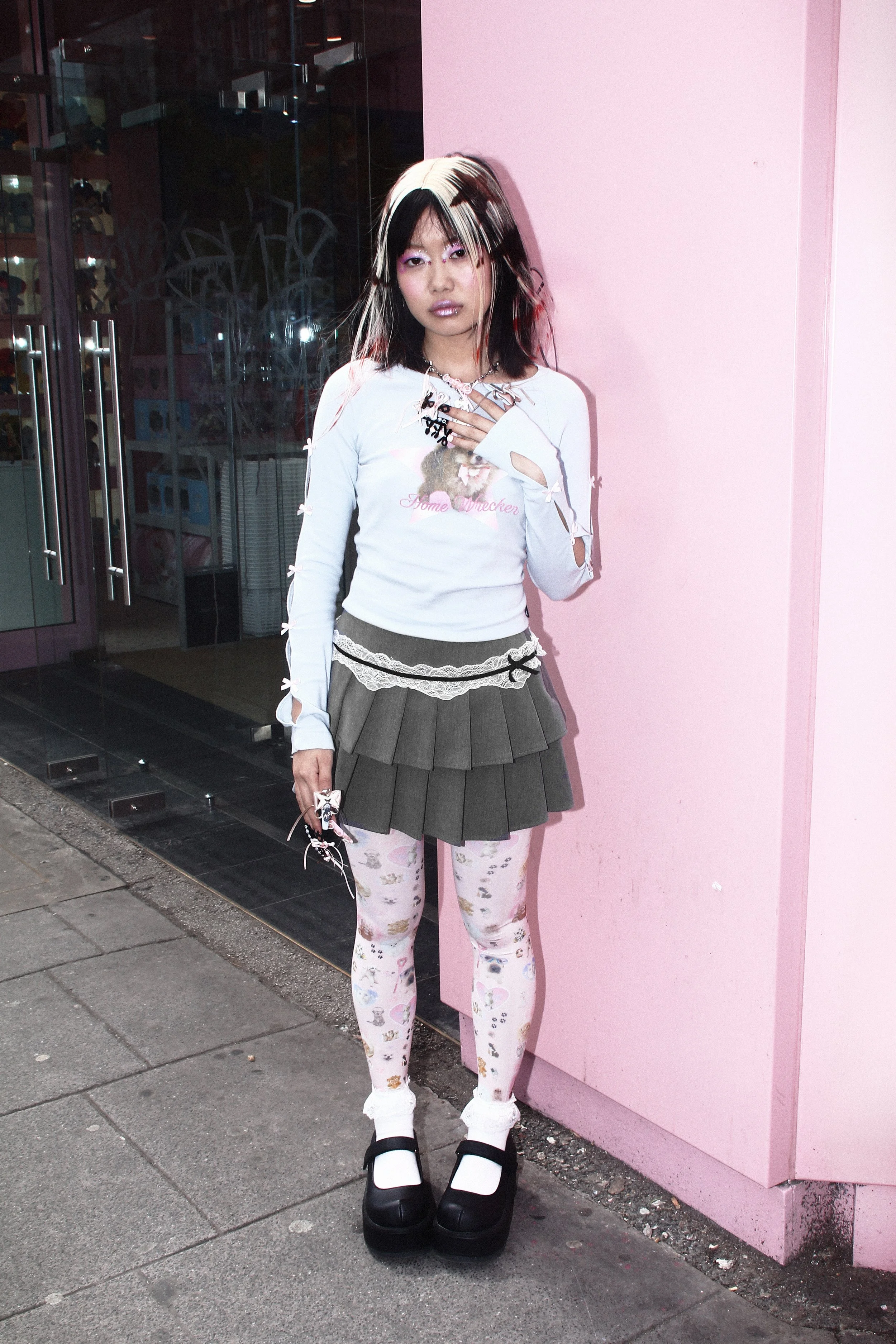 Full-body editorial image of a model in a pastel blue fitted top, pleated mini skirt, printed tights and chunky platform Mary Jane shoes standing against a pink storefront wall in Camden.