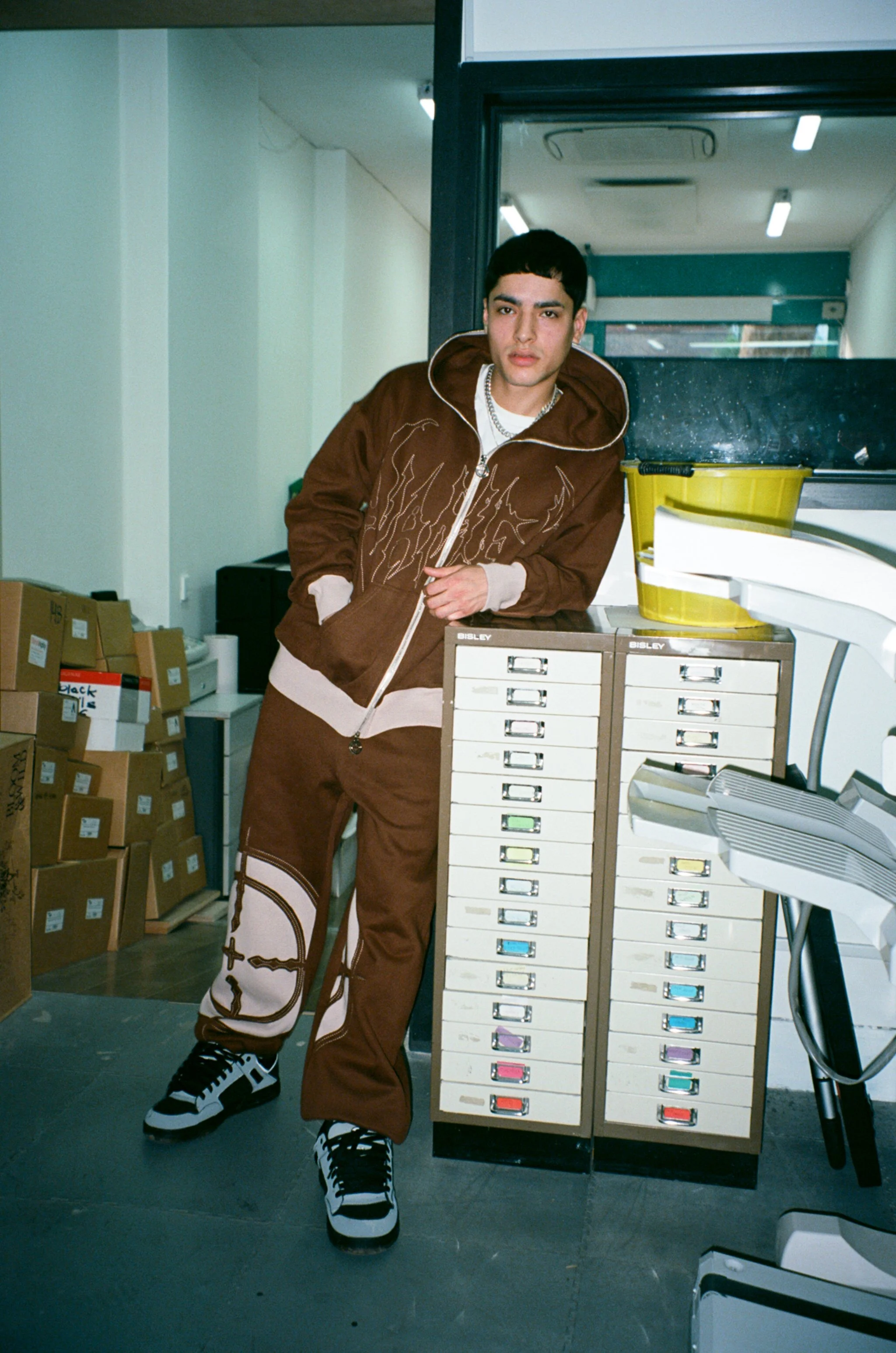 Model wearing a brown NAMED Collective “Classified” tracksuit with embroidered graphics, photographed in an office-style space with filing drawers, cardboard boxes and bright flash lighting.