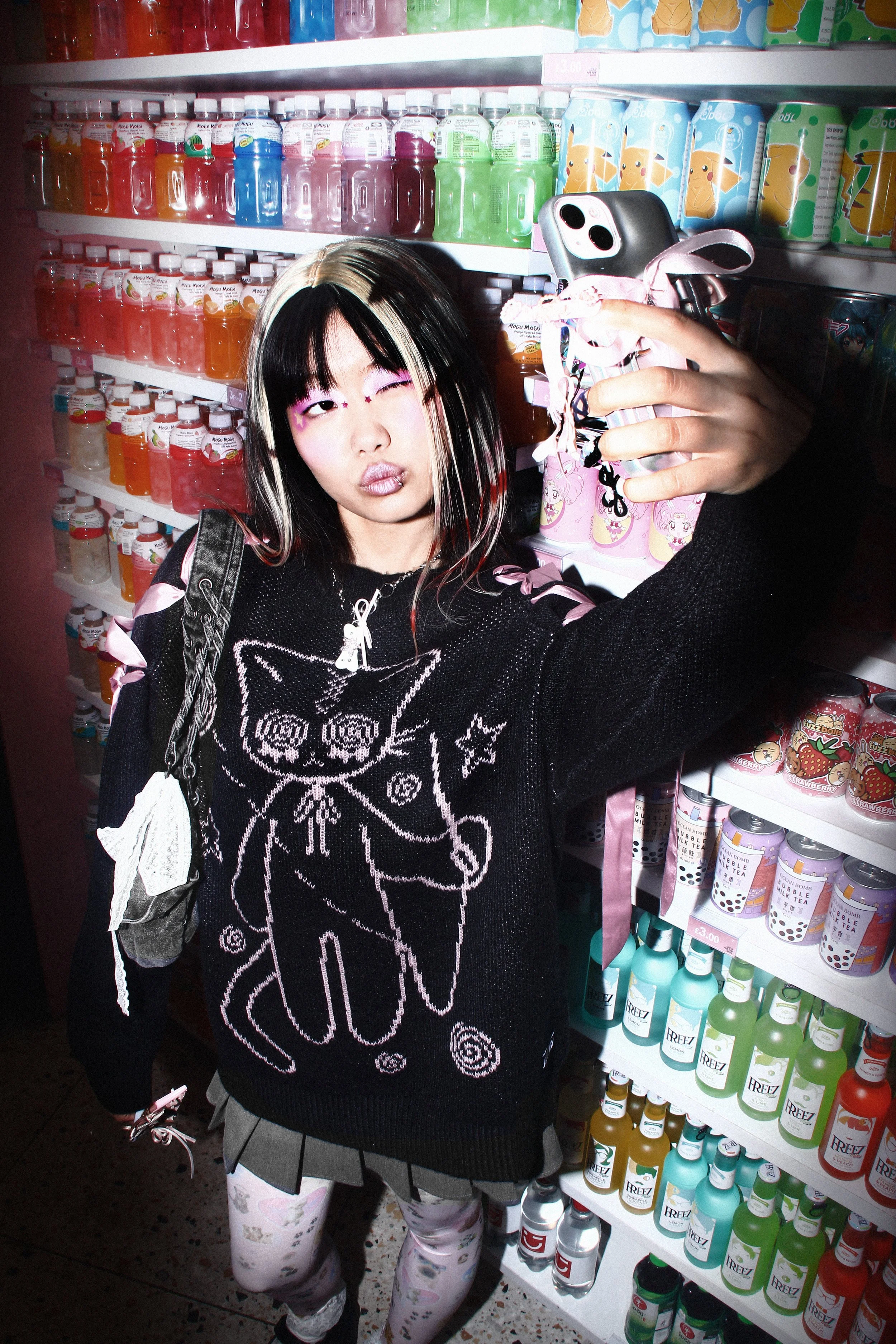 Streetwear-inspired campaign image of a model taking a photo inside a brightly coloured Japanese convenience store while wearing a black graphic knit sweater, pleated skirt and layered accessories for Minga London.